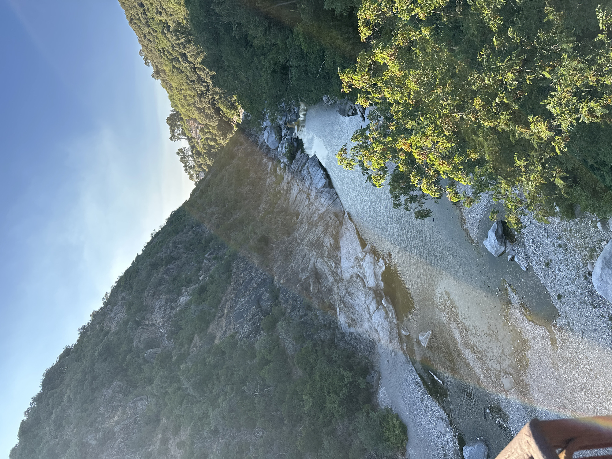 An aerial perspective captures a serene natural landscape. A river winds through a narrow valley, flanked by steep, tree-covered hillsides. The riverbed is characterized by smooth, grey stones and rocky outcrops, with a section of shallower water revealing submerged rocks and pebbles. Sunlight filters through the trees, creating dappled patterns on the water and ground. In the distance, nestled amongst the trees on the right hillside, are a few small buildings, suggesting a human presence in the otherwise wild environment. The sky above is a clear, bright blue. The foreground shows a sliver of a weathered, reddish-brown railing, possibly part of a bridge or viewpoint, from which the photograph was taken. The overall scene evokes a sense of tranquility and untouched natural beauty, indicative of a rural or mountainous region.