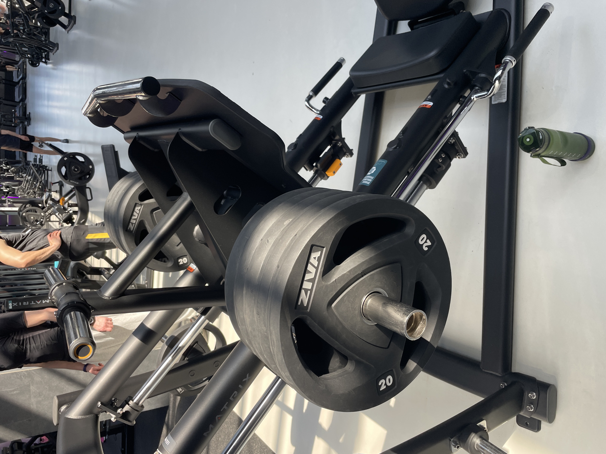 The image shows a leg press machine with "ZIVA" brand weight plates loaded on the weight horn in what appears to be a gym or fitness center. The machine is black and has "MATRIX" branding visible. There are other exercise machines and people in the background, which are slightly out of focus. The setting appears to be indoors with a white wall behind the leg press machine. A green water bottle is attached to the wall. I am unable to identify the city of Kazan, Russia from the image.