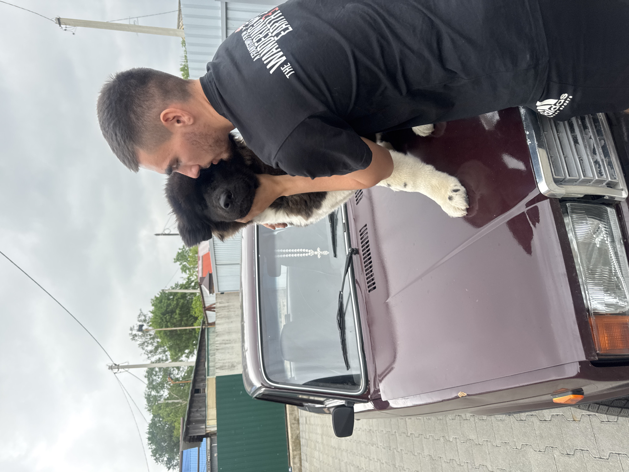 An adult male, with short dark hair, wearing a black t-shirt with white text that reads "THE WANDERING EARTH", is shown from the chest up, embracing a fluffy, black and white puppy. The man is leaning over the hood of a dark red, older model sedan, with his face close to the puppy's head. The puppy is positioned on the car's hood, with its paws resting on the metal surface. A string of white prayer beads with a cross charm hangs from the car's rearview mirror. The car is parked on a gray paver stone surface. In the background, there are fences, buildings, trees, and utility poles under a cloudy sky. The scene appears to be in an outdoor urban or suburban environment.