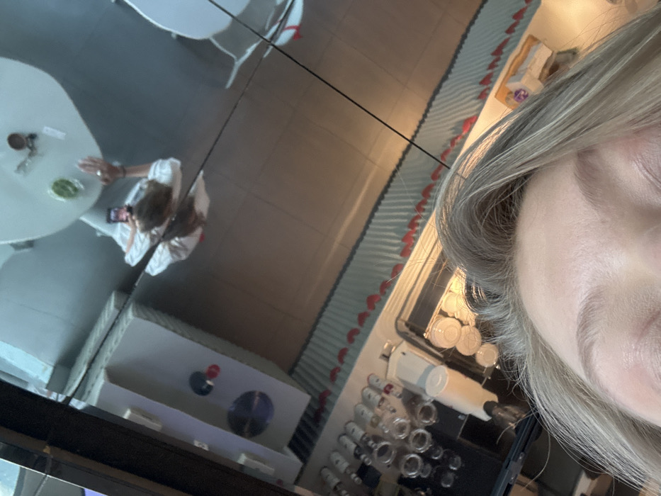 A woman's face dominates the right side of the frame, her gray hair partially obscuring the scene. The image reflects in a surface, showing a table with dishes and a person in a white shirt holding a phone. Also reflected is a cabinet with circular elements, along with rows of jars and kitchenware. The overall composition is fragmented and distorted due to the reflective surface.
