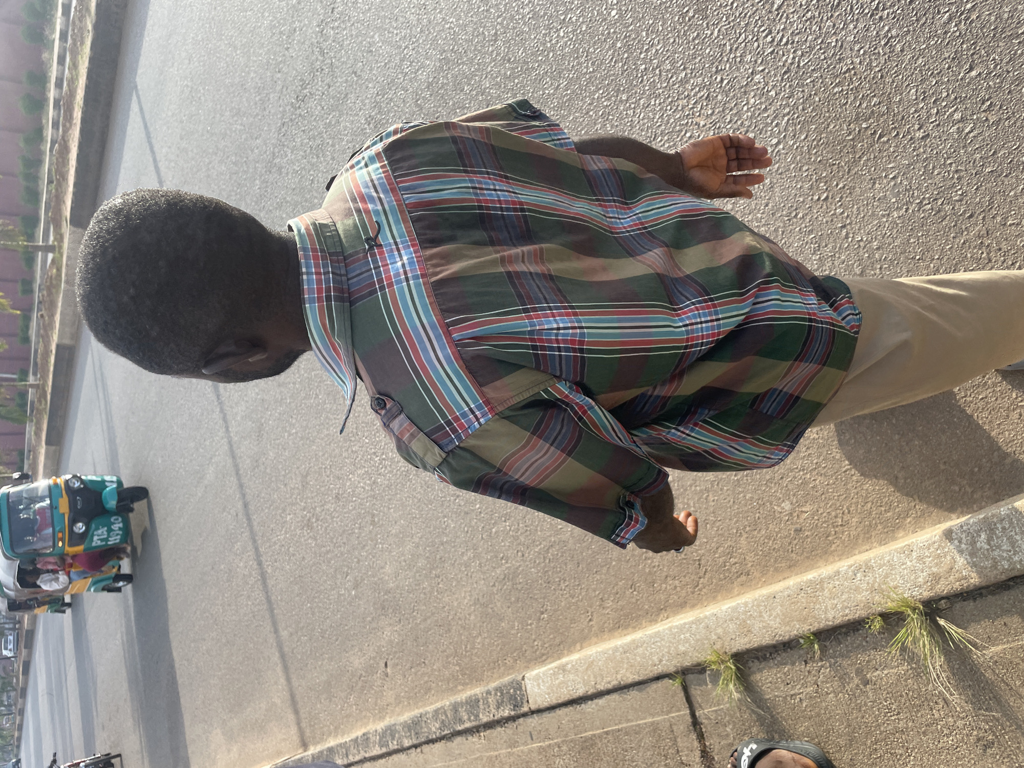 The image depicts a young Black male walking away from the camera on a paved road in Jos, Nigeria. He is wearing a plaid shirt with green, red, blue, and brown stripes, and khaki pants. The road is bordered by a concrete sidewalk with some grass growing through cracks. In the background, a three-wheeled vehicle, commonly known as a "Keke Napep" or "Maruwa," is visible on the road, carrying passengers. The scene suggests a typical urban environment in Jos, characterized by the presence of these local transportation vehicles and the general infrastructure of the city. The lighting indicates it is likely daytime, with the sun casting shadows.