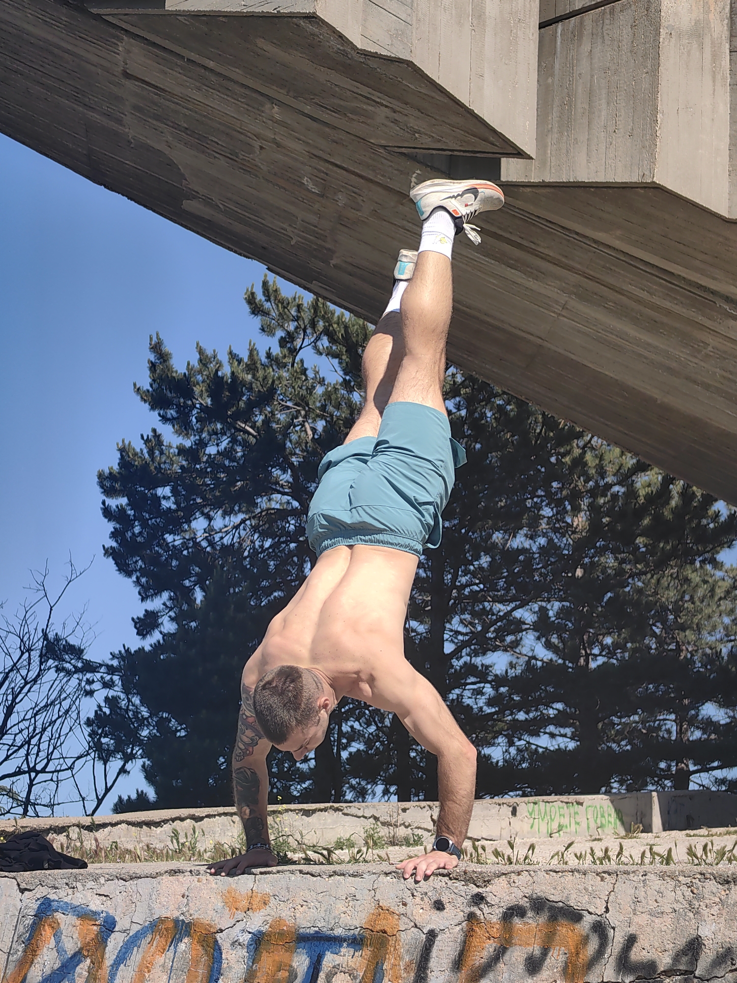 A man is performing a handstand outdoors on a weathered concrete wall. He is shirtless and wearing light blue shorts, white socks, and running shoes. He has tattoos on his left arm and is wearing a watch on his left wrist. His body is positioned beneath a large, angled concrete structure. Behind him are evergreen trees and a clear blue sky. The wall has graffiti markings. The location is the Monument of the Bulgarian-Soviet Friendship in Varna, Bulgaria.