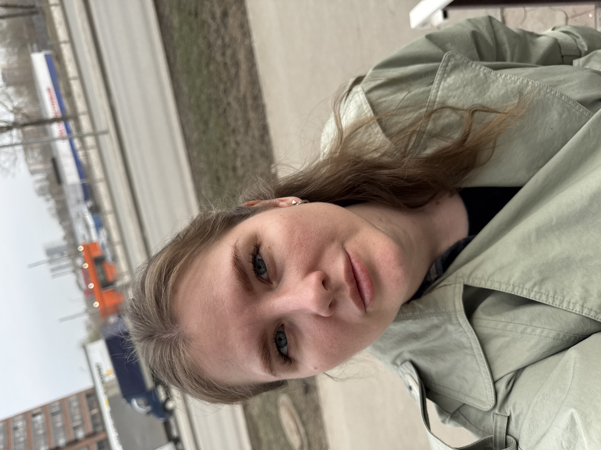 A woman is in the city of Perm, Russia, taking a selfie while sitting on a bench. A road with traffic, a building, and a median are visible in the background. The woman has light brown hair and is wearing a light green jacket. She has green eyes. She is likely sitting on a bench in a public area.