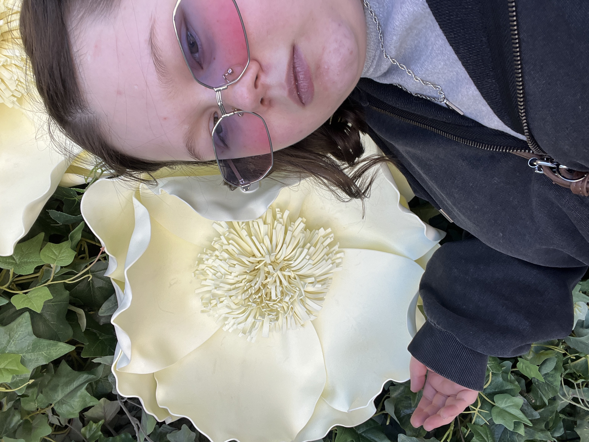 A young person is captured in a close-up, high-angle shot, lying down and looking towards the camera. Their head is tilted, and they wear large, silver-framed, pink-tinted sunglasses that obscure their eyes. They have dark hair and light skin, with some minor blemishes visible on their forehead and cheeks. Their lips are slightly parted and appear to have a neutral-toned lip product.
The person is dressed in a dark, possibly black, zip-up jacket worn over a grey inner garment, which could be a turtleneck or hoodie. A silver chain necklace is visible under the jacket's collar. One hand, with light skin, rests gently on the green ivy leaves that cover the ground.
The setting features a large, artificial flower in a cream or light yellow color, with ruffled petals and a distinct, fringed center resembling stamens. This flower is positioned prominently near the person's head. The ground is covered with a dense layer of green ivy. The lighting is bright and even, indicating daytime. The person's expression is neutral, and no specific emotions are strongly conveyed. There is no visible text in the image. The location context for this scene is Yekaterinburg, Russia.
