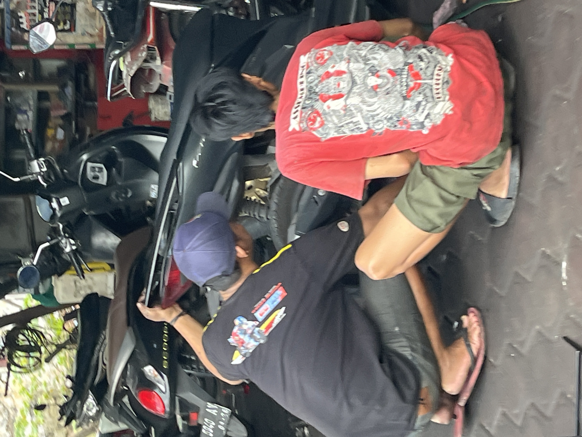 The image shows two individuals, likely mechanics, working on motorcycles in what appears to be a repair shop in Kepatihan, Indonesia. One person, wearing a black t-shirt with a graphic print and a blue baseball cap, is seen from behind and the side, reaching towards the rear of a black scooter. The other person, wearing a red t-shirt with a detailed graphic print and olive green shorts, is also seen from behind, seated and looking towards the motorcycle. They are both kneeling or crouching, engaged in repair work. The environment is cluttered with motorcycles and various shop equipment. The floor is dark and textured, possibly a rubber mat. Various parts of motorcycles, including seats, handlebars, and license plates, are visible. The overall impression is of a busy, functional workspace dedicated to motorcycle maintenance.