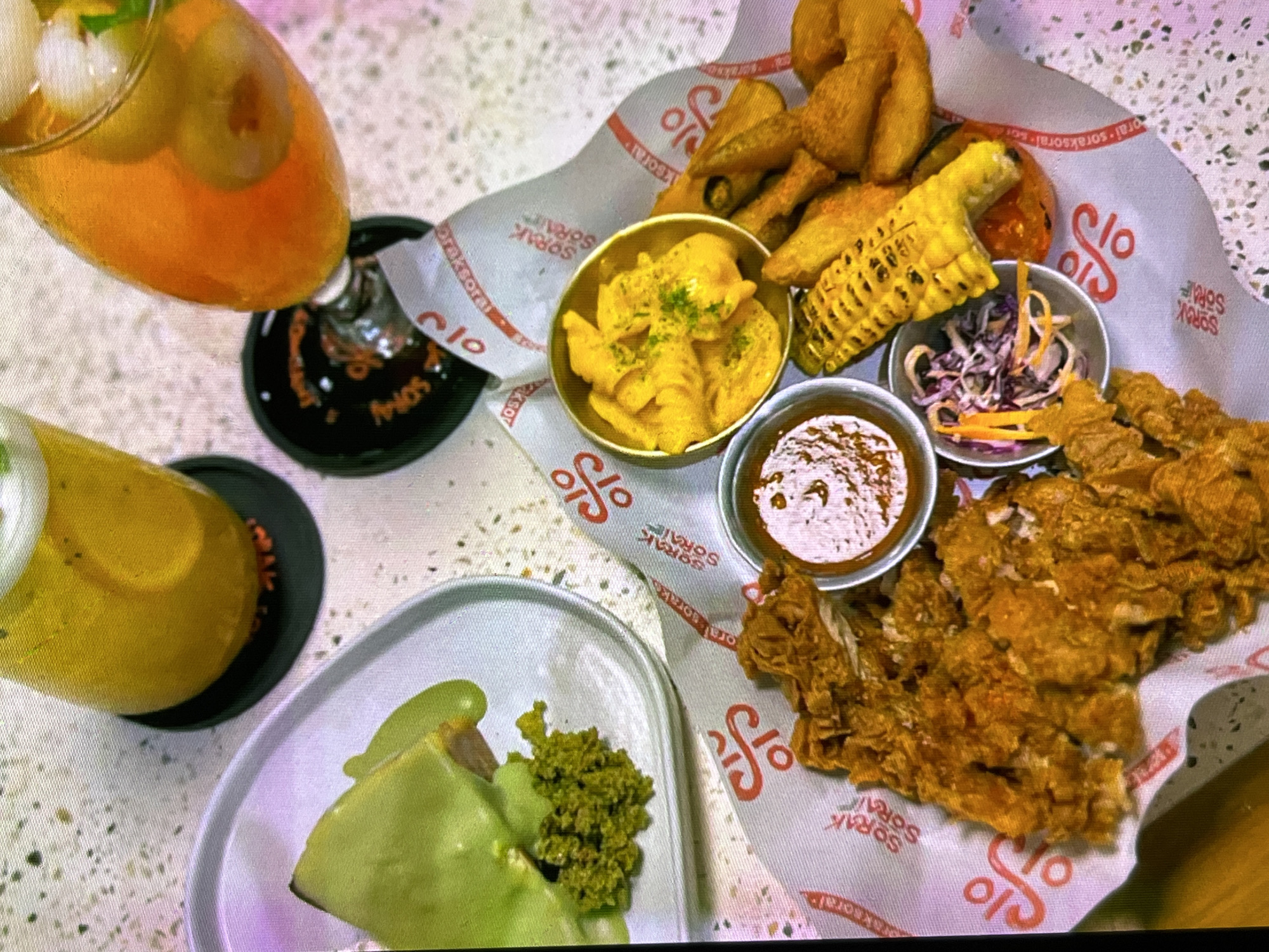 A top-down, medium shot captures a table at "Sora Sorai" in Cadek, Indonesia, featuring a collection of food and drinks. In the foreground, a large serving of golden-brown fried chicken rests on a patterned paper liner, accompanied by a small bowl of dipping sauce and a side of coleslaw. Adjacent to this is a grilled corn on the cob and several potato wedges. To the left, a slice of cake, seemingly topped with a vibrant green sauce and a sprinkle of green crumbs, sits on a white plate. Two drinks are visible: one on the left is a tall glass filled with a yellow beverage and ice, placed on a black coaster; the other, on the right, is a stemmed glass containing an orange-hued drink with ice and fruit slices, also on a black coaster. The table surface is a speckled, light-colored material. The "Sora Sorai" logo appears repeatedly on the paper liner.
