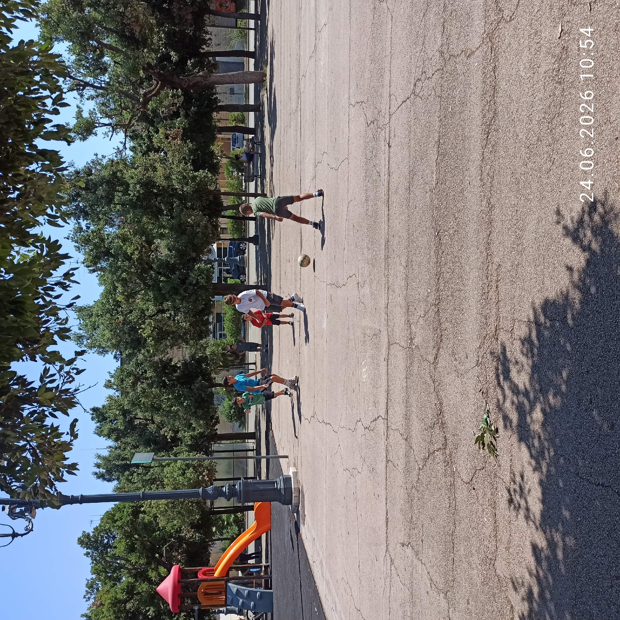 The image captures a daytime scene at an outdoor public space, possibly a park or playground, in Bitonto, Italy. Several young people are engaged in a game of soccer on a paved area with visible cracks. The sun is shining brightly, casting distinct shadows of trees and people onto the ground, indicating it is likely midday. Lush green trees line the background, suggesting a pleasant, possibly warm, weather condition.
In the foreground, a playground structure with colorful slides, including an orange one, is partially visible. The primary focus is on the group of individuals playing soccer. One person in a green t-shirt and grey shorts is kicking the ball. Another person in a white t-shirt and dark shorts appears to be observing or participating. Further down the paved area, two other individuals in blue and green t-shirts are also present, possibly waiting their turn or engaged in other activities related to the game.
The overall atmosphere seems casual and active, with the individuals appearing focused on their game. The presence of the playground suggests it's a family-friendly location. A timestamp "24.06.2026 10:54" is visible in the top right corner, indicating the date and time the photo was taken. The scene is vibrant, with the bright sunlight and the activity of the young people contributing to a lively ambiance.