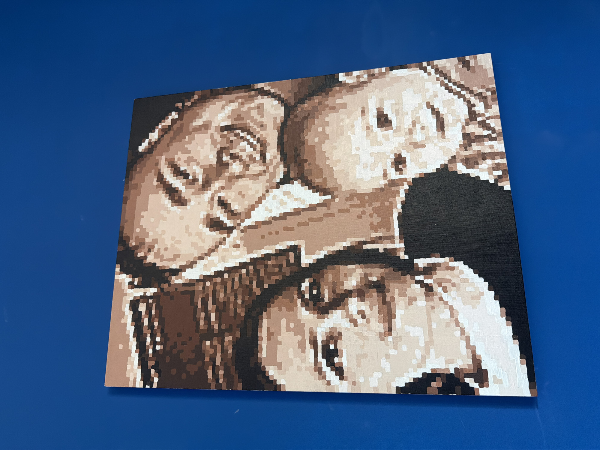 An intricately detailed pixel art painting is prominently displayed diagonally against a plain, vibrant blue background. The artwork, appearing to be on a canvas, features three distinct human faces rendered in a limited, monochromatic palette of various shades of brown, beige, white, and black.
In the upper left quadrant of the painting, a face with a wide, joyful smile is visible, characterized by discernible eyes, nose, and an open mouth revealing teeth. To its right, a second face, possibly that of a child due to softer features, looks intently towards the right with wide, expressive eyes and an open mouth. The third face is situated in the lower middle and right, mostly visible from the nose upwards, with open eyes and a slightly parted mouth, gazing slightly upwards and to the right. The faces within the artwork are also tilted, aligning with the diagonal orientation of the canvas itself.
The artwork's pixelated style gives it a distinct, blocky appearance, with each color segment clearly defined. The texture of the canvas surface is subtly visible. The solid blue background contrasts sharply with the detailed sepia tones of the painting, highlighting the artwork. No visible text is present. The lighting is bright, clearly illuminating both the painting and the blue surface it rests upon. The location is Chaikovsky, Russia.