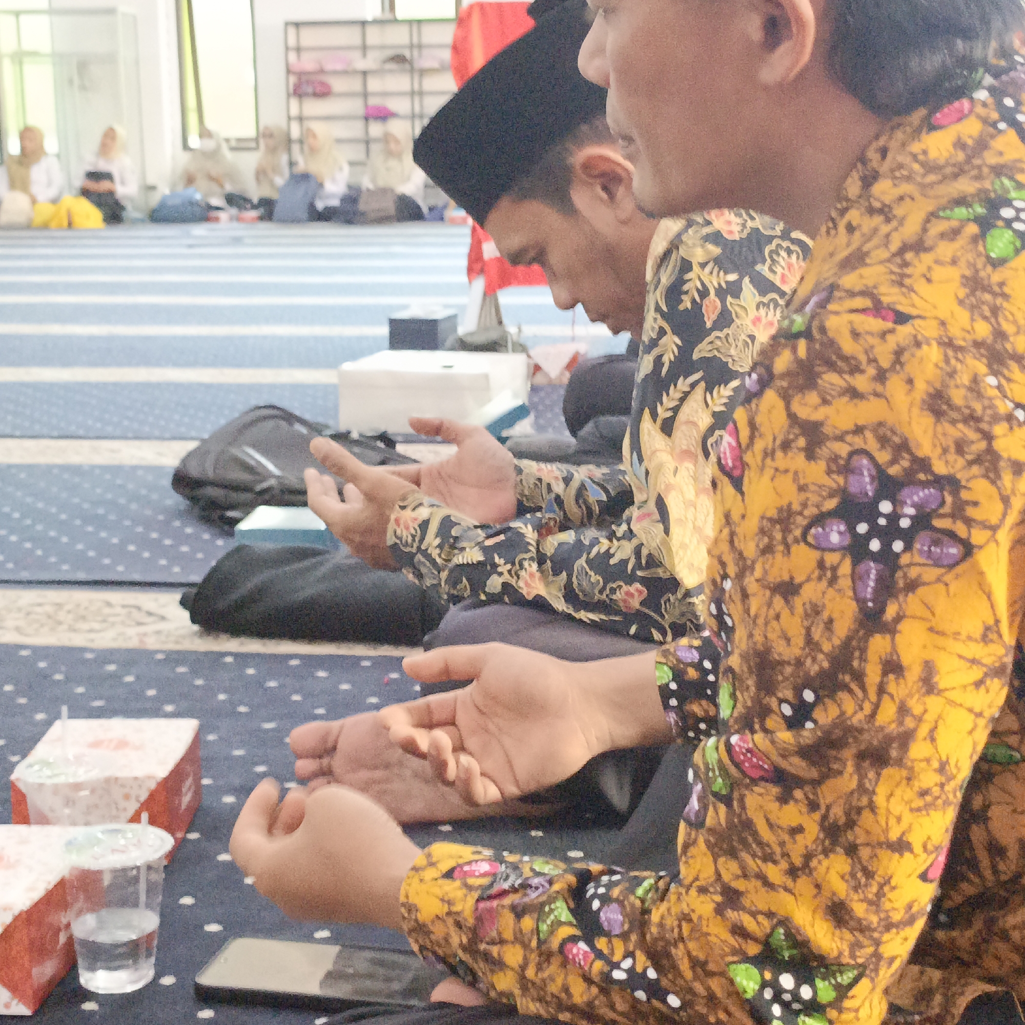 The image shows two young men sitting on a blue and white striped carpeted floor. Both are wearing traditional Indonesian batik shirts, one predominantly yellow with brown and purple floral patterns, the other dark blue with lighter floral patterns. The man on the left wears a black cap. Their hands are held open, palms up, in a gesture of prayer or supplication. In the background, other individuals, mostly women wearing headscarves, are also seated on the floor, blurred. Behind them, shelves holding various items are visible. Several red and white food boxes and a plastic cup of water are in the foreground. A smartphone is also visible on the floor. The setting appears to be an indoor space, possibly a mosque or a community hall in Salatiga, Indonesia, given the presence of prayer mats and the cultural attire.