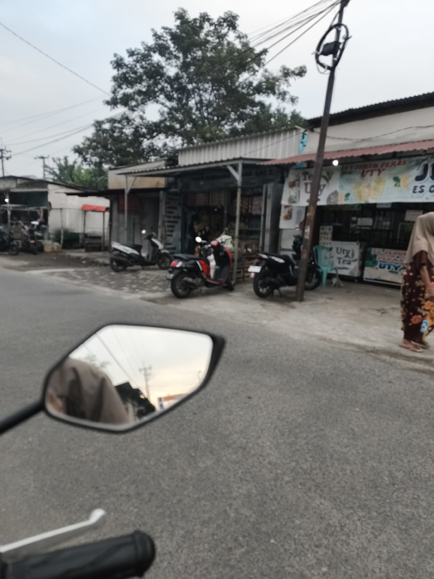 A street-level, slightly blurred photograph depicts a row of small shops lining a paved road in Sidorejo, Indonesia. The shops appear to be typical Indonesian roadside businesses, with awnings and open fronts displaying goods. Several motorcycles are parked or displayed in front of the shops. One red and black scooter is prominent on the right, and a white and black scooter is parked further back. A woman wearing a hijab and a patterned dress walks away from the shops on the sidewalk. In the foreground, a motorcycle's rearview mirror is visible, reflecting the sky and part of the scene. The overall atmosphere suggests a commercial area during daylight, possibly late afternoon or early morning given the lighting. A prominent utility pole with many wires stands near the shops.