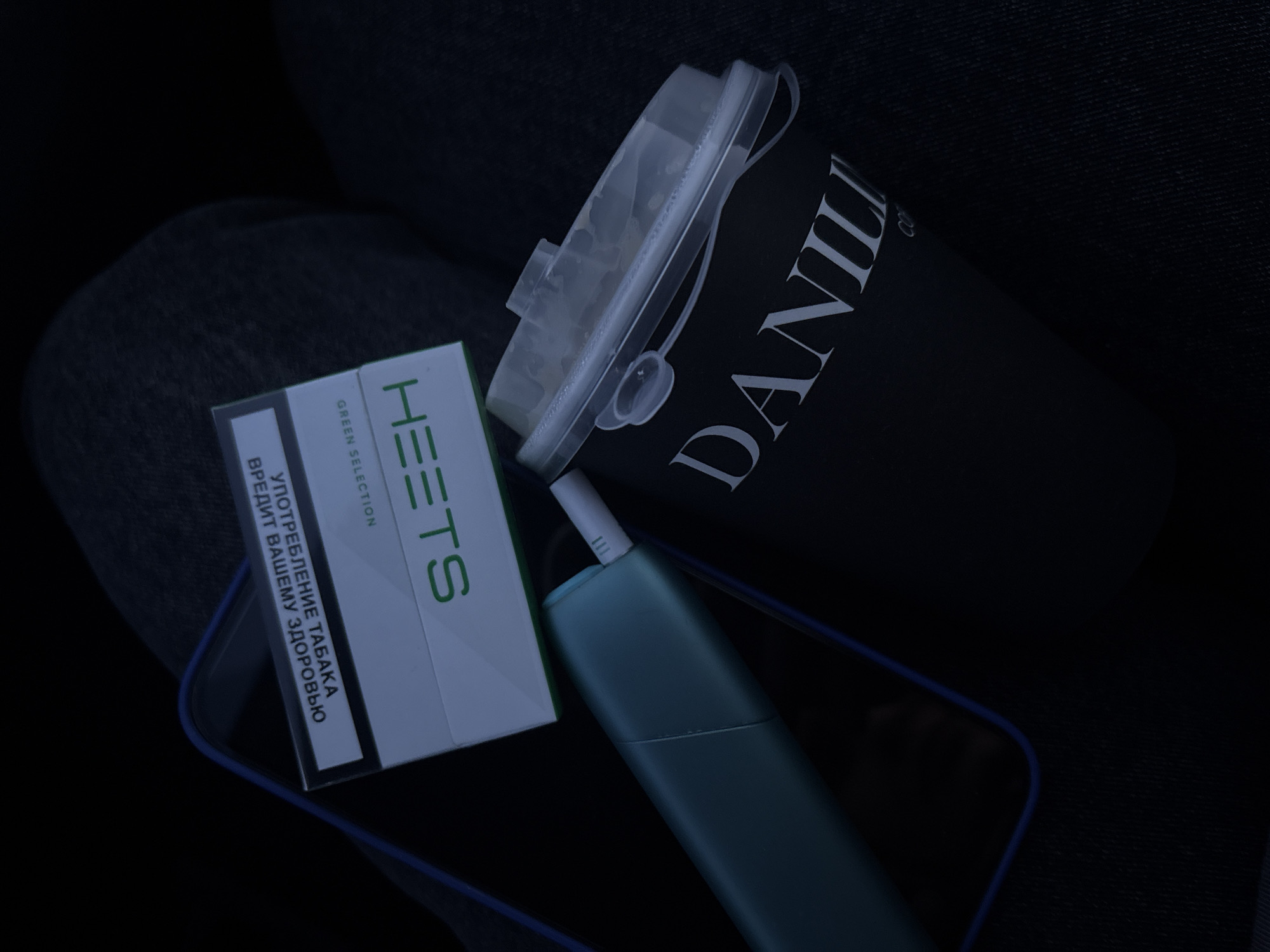 The image presents an overhead view of personal items arranged on a dark, textured fabric surface, likely a lap or seat, within the city of Chaikovsky, Russia. A white and green pack of HEETS 'GREEN SELECTION' tobacco sticks, bearing the Russian health warning "УПОТРЕБЛЕНИЕ ТАБАКА ВРЕДИТ ВАШЕМУ ЗДОРОВЬЮ" (Tobacco use harms your health), is positioned centrally. Beneath this pack, a smartphone with a blue-edged case is partially visible. To the right and below the tobacco pack, a teal-colored IQOS tobacco heating device is depicted with a white tobacco stick inserted into its holder. Further to the right, a black disposable coffee cup, branded "DANILI COF" in white lettering, is present, topped with a clear plastic sip-lid. The scene is dimly lit.