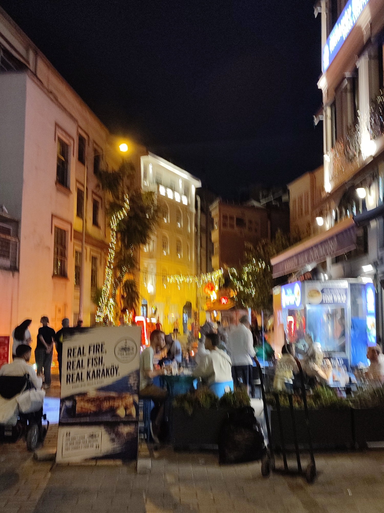 The image depicts a nighttime scene in the city of Beyoğlu, Turkey, likely in the Karaköy district, known for its vibrant street life and eateries. The perspective is from street level, looking down a narrow street lined with buildings on either side. The architecture suggests an urban environment with multiple stories, some adorned with decorative lights. A prominent sign in the foreground advertises "REAL FIRE, REAL FISH, REAL KARAKÖY," indicating a food establishment, possibly a fish restaurant. Several people are visible, some standing and conversing, while others are seated at tables, presumably dining or socializing. One individual is using a wheelchair. The atmosphere appears lively and bustling, with the glow of artificial lights reflecting off the wet pavement, suggesting recent rain or a damp environment. The overall impression is one of an active urban night scene centered around food and social gathering.