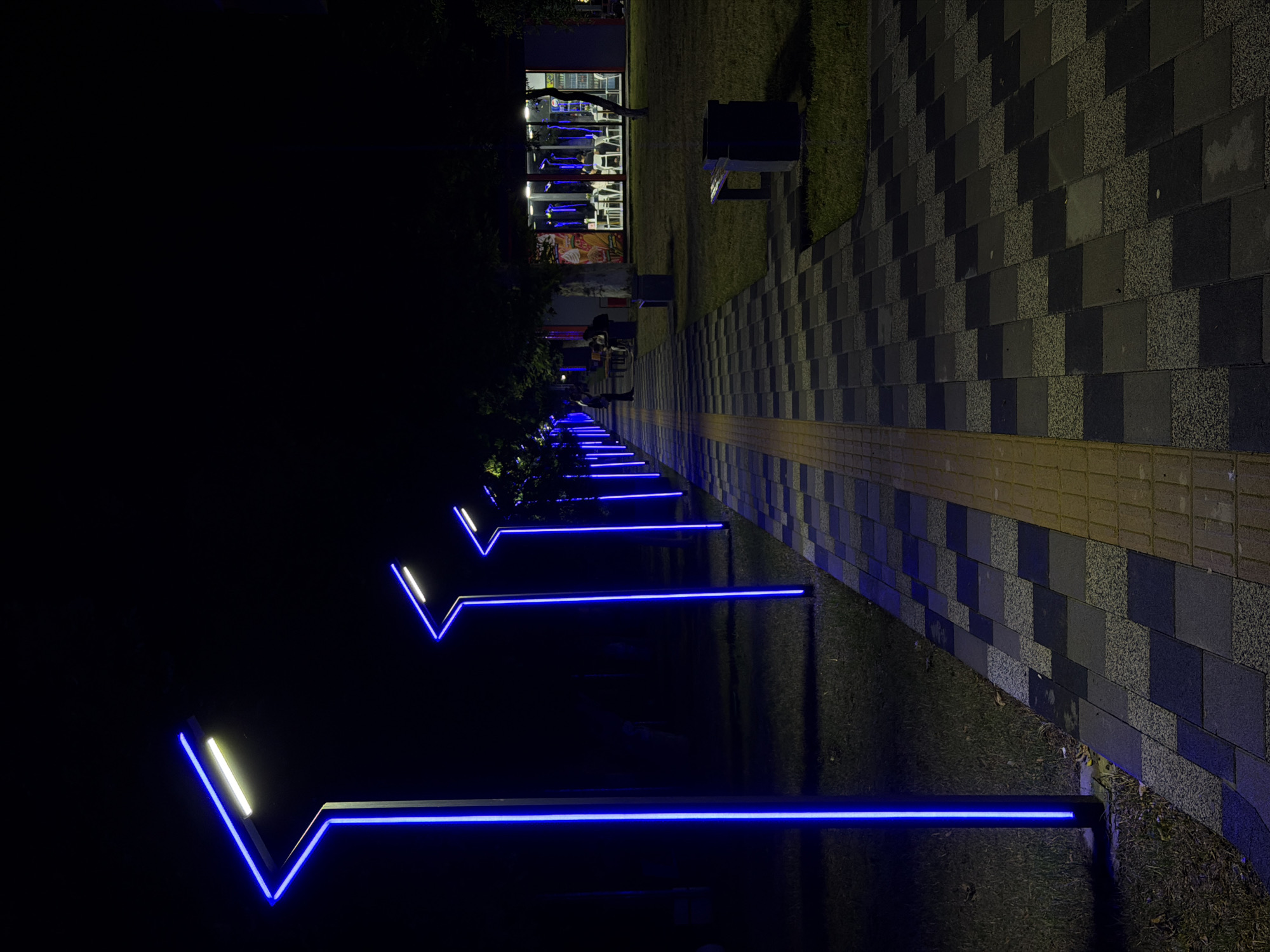 The image shows a pedestrian walkway at night, illuminated by modern blue LED streetlights. The walkway is paved with dark gray and black checkered tiles, with a textured yellow tactile paving strip along its edge. On the left side of the walkway, there is a patch of grass and a row of the distinctive blue-lit streetlights. In the background, on the right side, a modern building with large glass windows is visible. Inside the building, tables and chairs are set up, and several people are seated, suggesting it is a cafe or restaurant. The overall scene depicts a well-lit, contemporary urban public space. This scene is located in Chirchik, Uzbekistan.