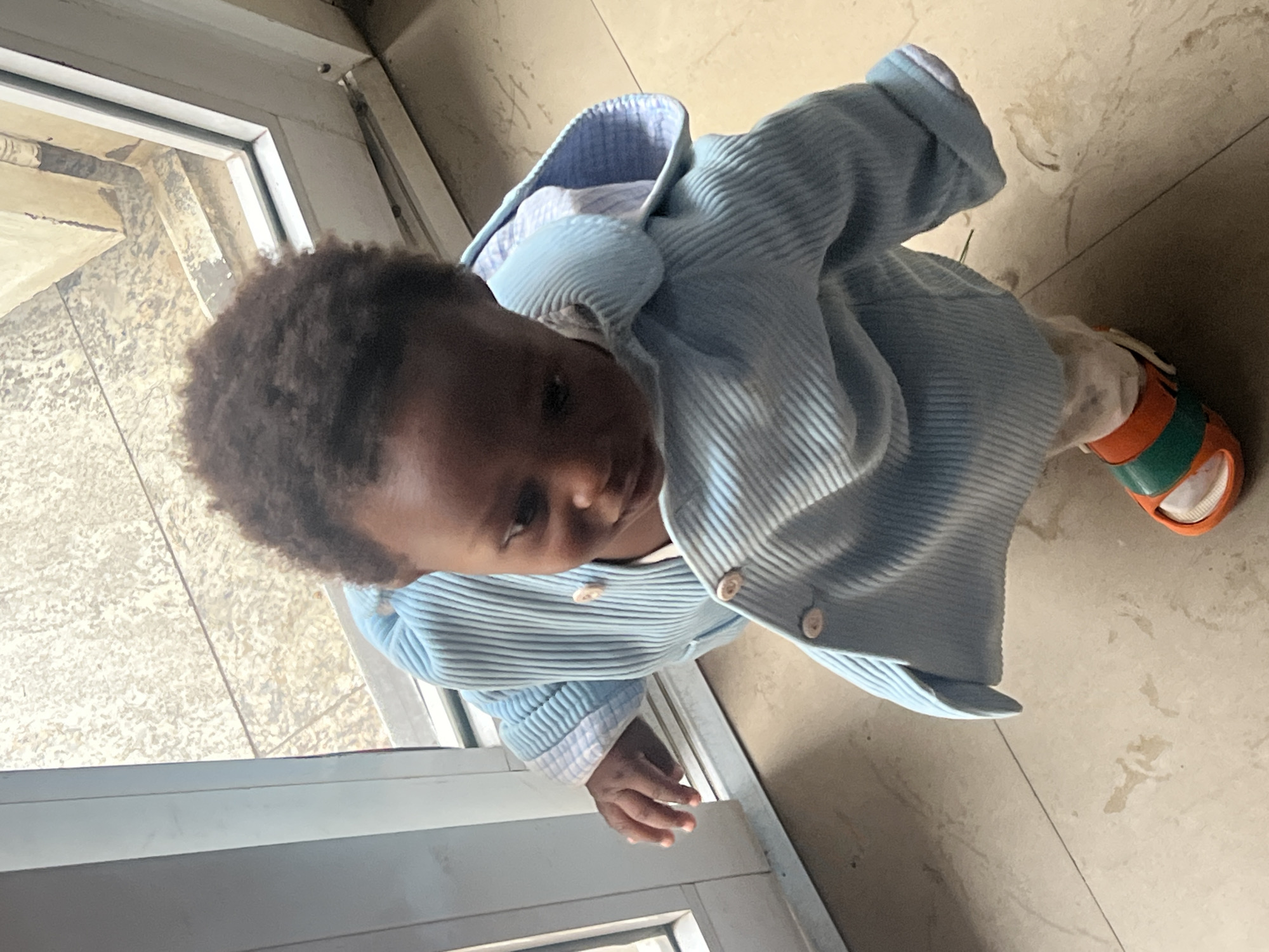 A child stands indoors near a window. The child wears a light blue, corduroy-like jacket, a white shirt, and orange and green sandals. The floor is tiled, and the window frame is metal. The location appears to be Gura Top, Nigeria.
