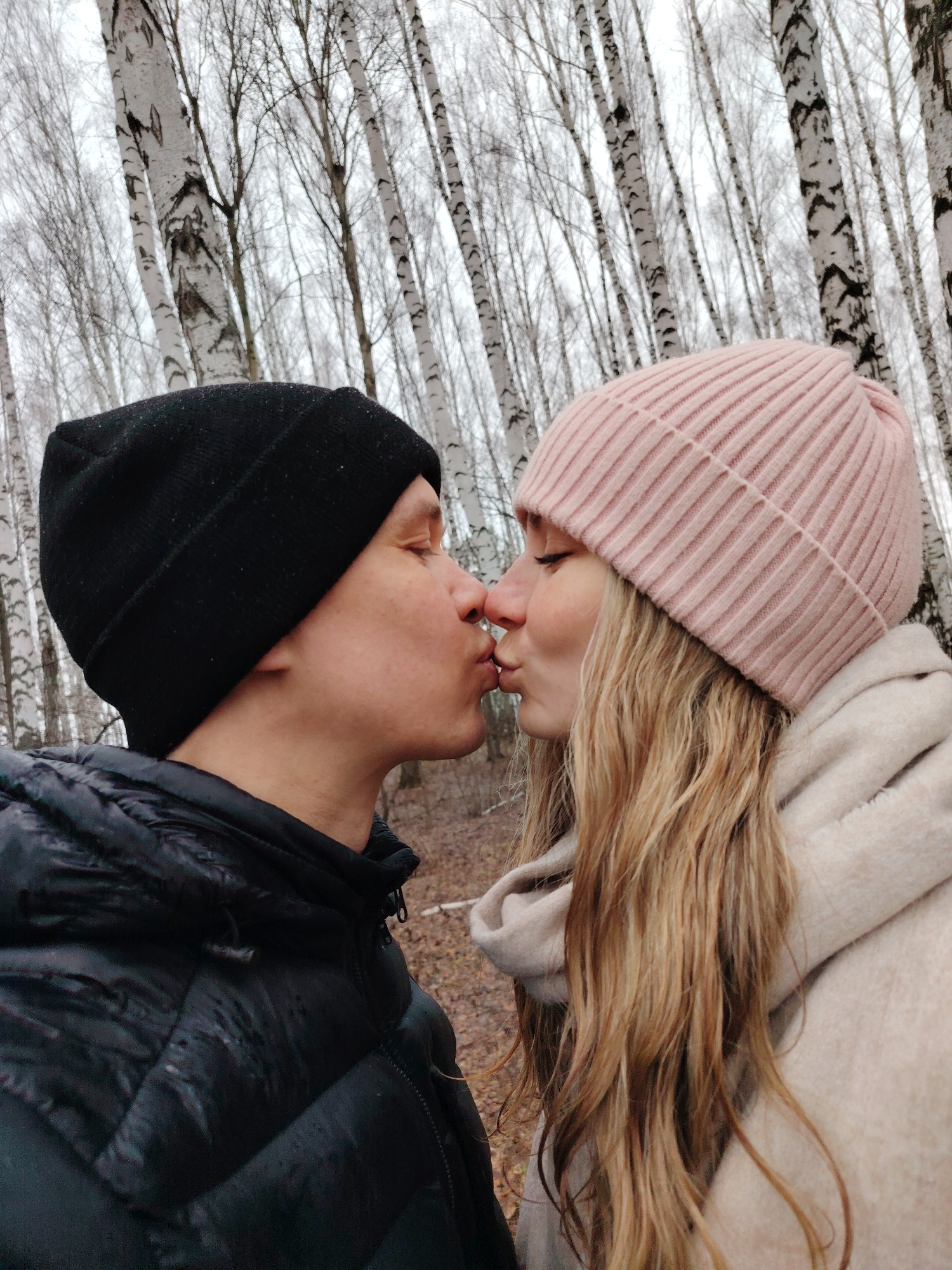 A couple is kissing in a birch forest. The man is wearing a black beanie and a black puffer jacket. The woman has long blonde hair and is wearing a pink beanie and a light-colored hooded jacket. They are surrounded by birch trees with white trunks and bare branches. The location is Chaikovsky, Russia.