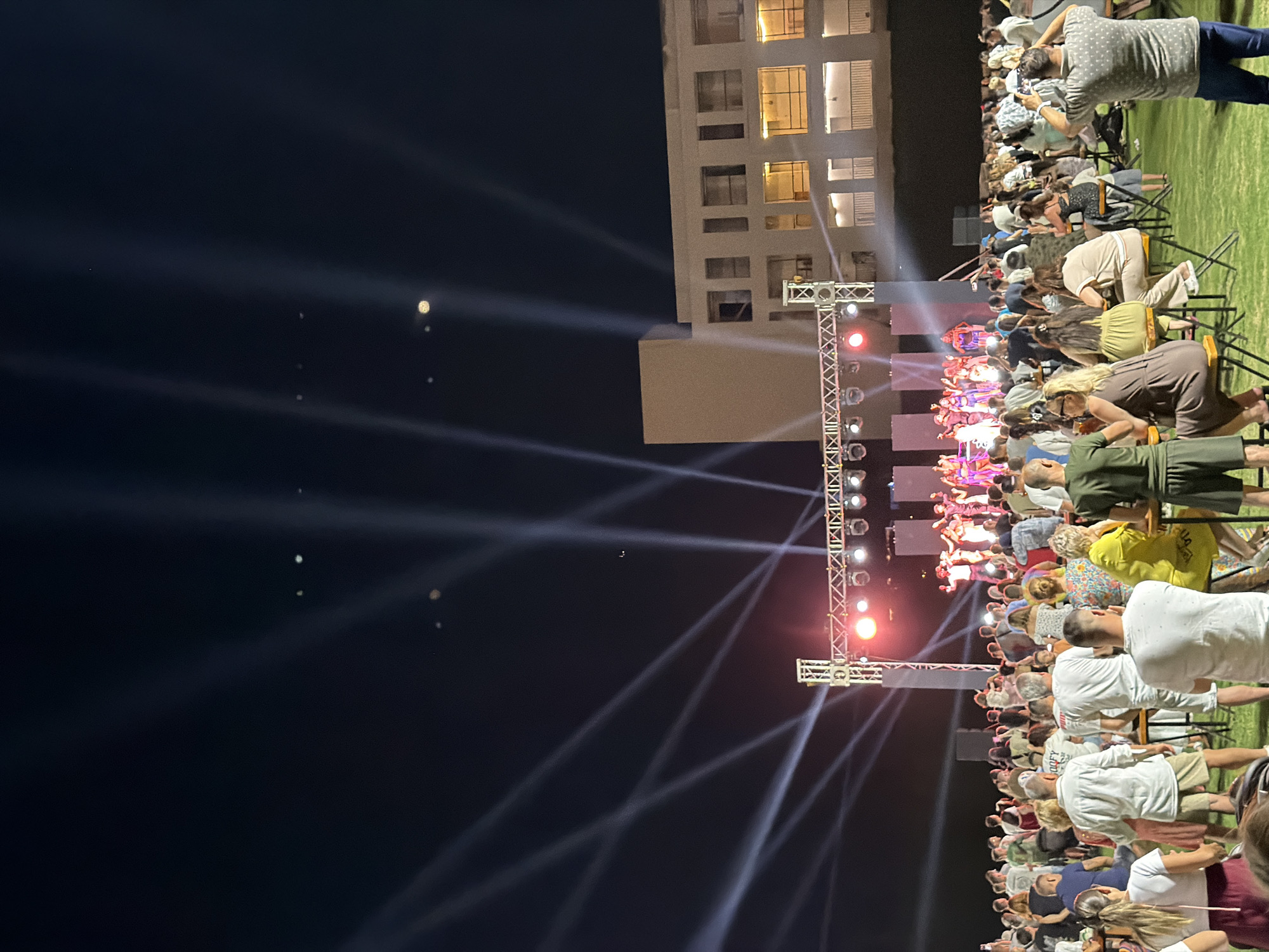 In Sharm El Sheikh, Egypt, a nighttime outdoor event is in progress. A stage is set up with bright lights illuminating performers, who appear to be dancers, dressed in colorful costumes. Behind the stage, a modern building with lit windows stands against the dark sky, crisscrossed by dramatic spotlights. A large crowd of people are gathered in front of the stage, some seated at tables and others standing. The ground is covered in green grass. Many attendees are facing the stage, suggesting they are watching the performance. One person in the foreground is taking a photograph of the event with their phone. The atmosphere is lively and festive, typical of a resort town event.