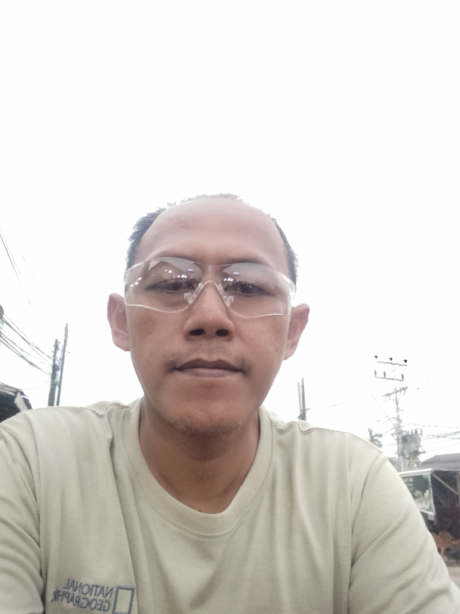A middle-aged man with a shaved head and light stubble on his chin is captured in a close-up, frontal portrait. He is wearing clear safety glasses and a light tan t-shirt with a logo that appears to say "NATIONAL GEOGRAPHIC" in reverse. The background is washed out and bright, suggesting an overcast sky. To the left of the man, power lines and poles are visible, indicative of an outdoor urban or semi-urban environment. To the right, there are glimpses of structures, possibly shacks or market stalls, and vegetation. The image appears to be taken in Arang Limbung, Indonesia, as indicated by contextual information.