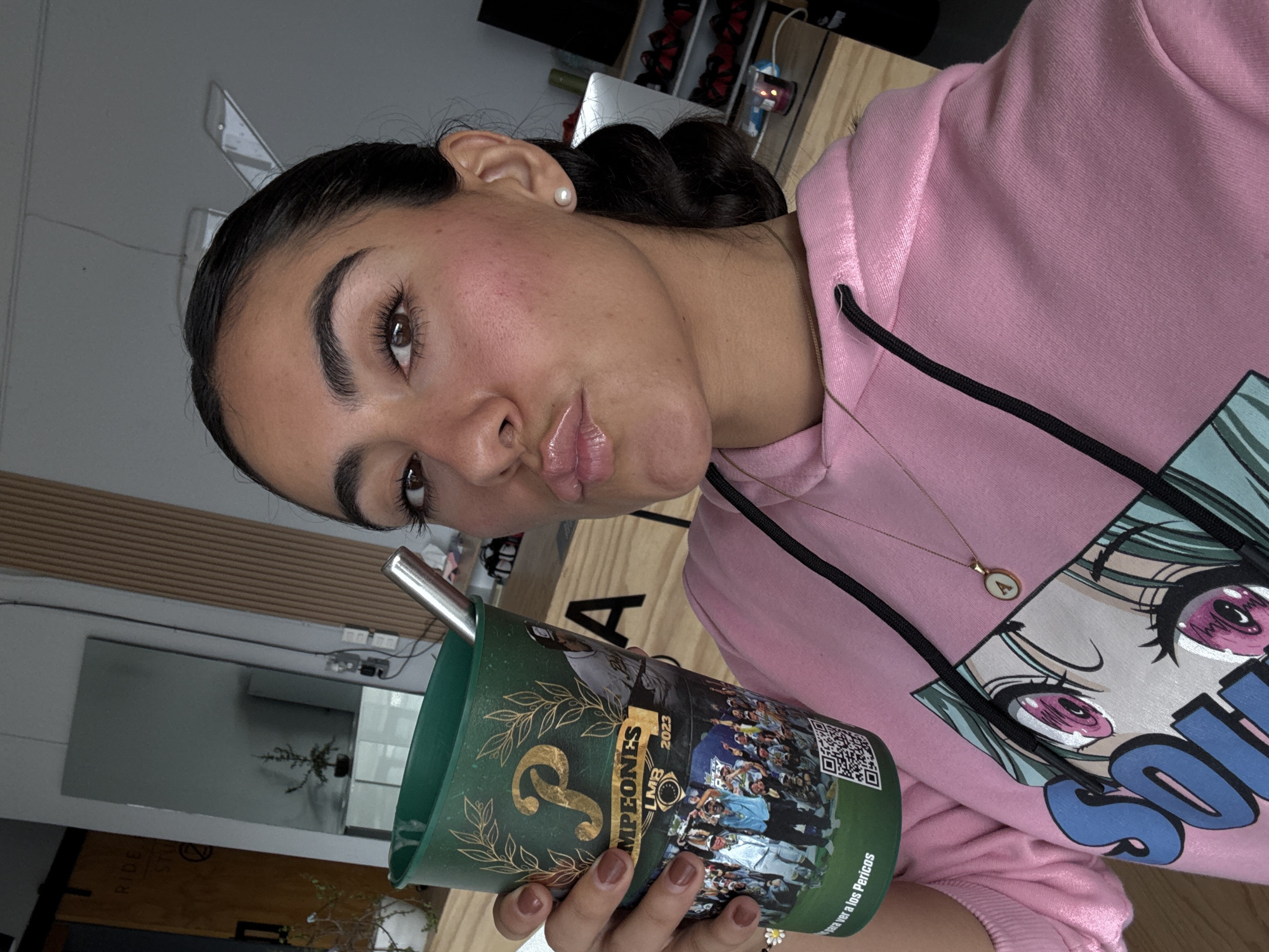 A medium, canted, eye-level shot shows a young woman in Puebla, Mexico, holding a green Pericos de Puebla Campeones LMB 2023 commemorative cup with a silver straw.
The woman has brown eyes, arched eyebrows, and glossy lips, wearing a pink hoodie with an anime-style character print and a gold necklace with a circular pendant. She has dark hair styled with an ear pearl and brown painted fingernails.
The background shows a room interior with a mirror, a wooden countertop and shelving unit, and a glimpse of outdoor greenery.