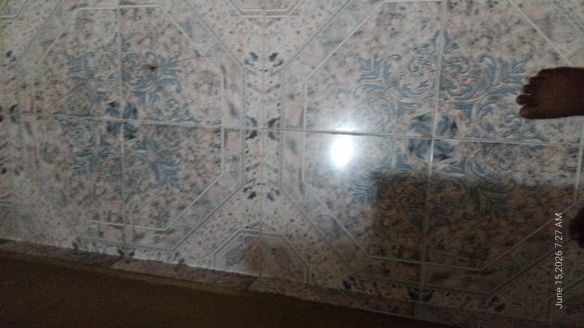 The image shows a tiled floor with an octagonal pattern. The tiles are a mix of beige, brown, and gray with blue floral designs. The lighting suggests an indoor environment, possibly a home or an office. In the bottom right corner of the image, a foot is visible, suggesting a person is standing on the floor. The timestamp "June 15, 2026 7:27 AM" is also visible in the lower left corner. The location in Howayi, Nigeria is not discernible from the image alone.