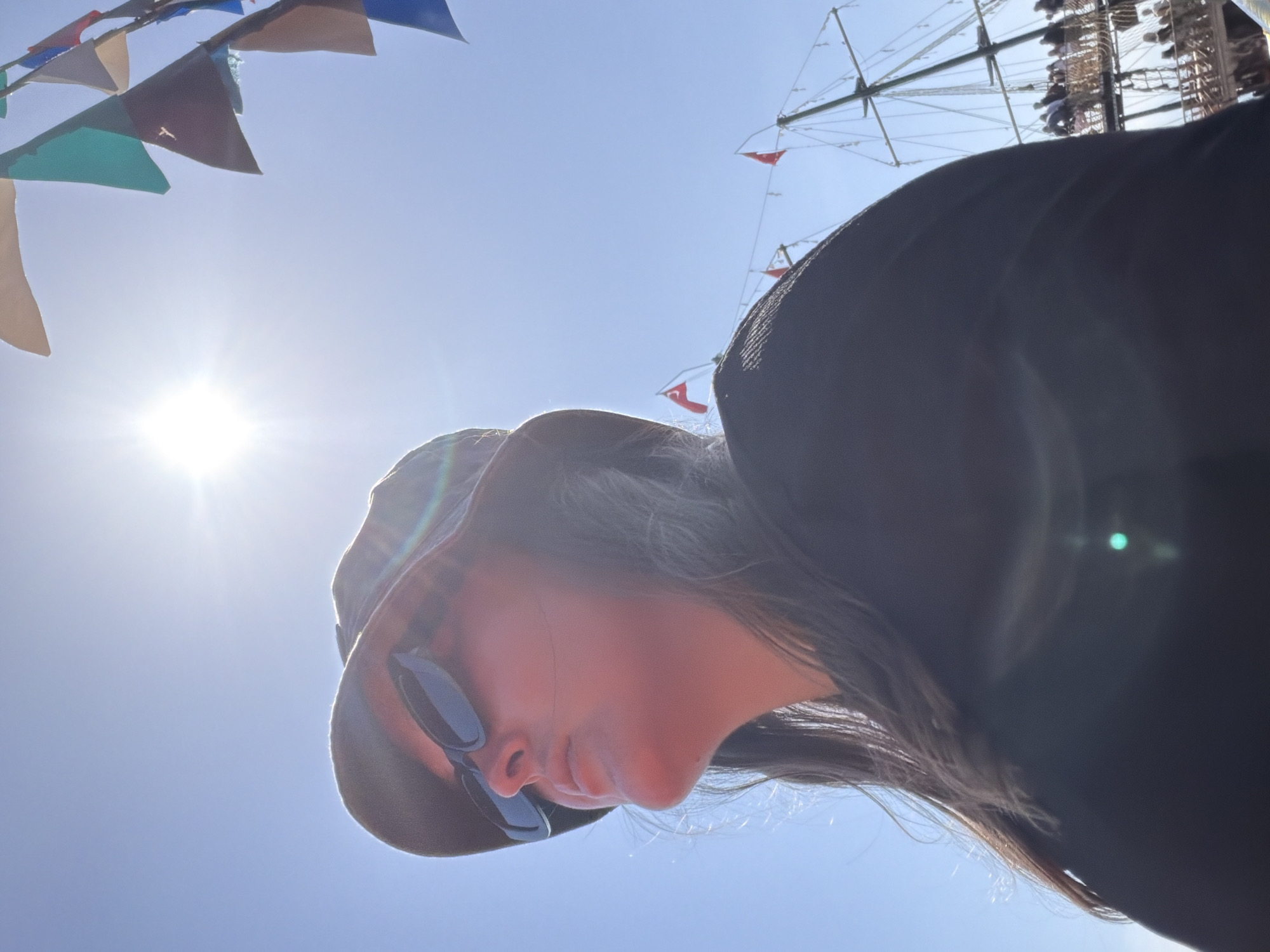 The image, taken from a low-angle perspective, looks upwards at a person and the sky above. A person, seen in profile, occupies the right side of the frame, wearing a dark brimmed hat and dark sunglasses. Their face is partially obscured by these accessories, and dark hair is visible beneath the hat. The bright sun, positioned in the upper left, causes a prominent lens flare.
Against a clear blue sky, colorful triangular flags (bunting) are strung across the top left corner, adding a festive touch. In the upper right background, the intricate rigging and mast of a tall ship are visible. Several small red pennants, each featuring a white crescent moon and star – the national emblem of Turkey – are attached to the ship's rigging, indicating the location as Alanya, Turkey, a coastal city.
The scene conveys a bright and sunny outdoor day, likely at a harbor or waterfront, with clear weather and abundant sunshine. The person appears to be looking slightly downwards or straight ahead from the camera's upward vantage point. No discernible text is visible in the image.