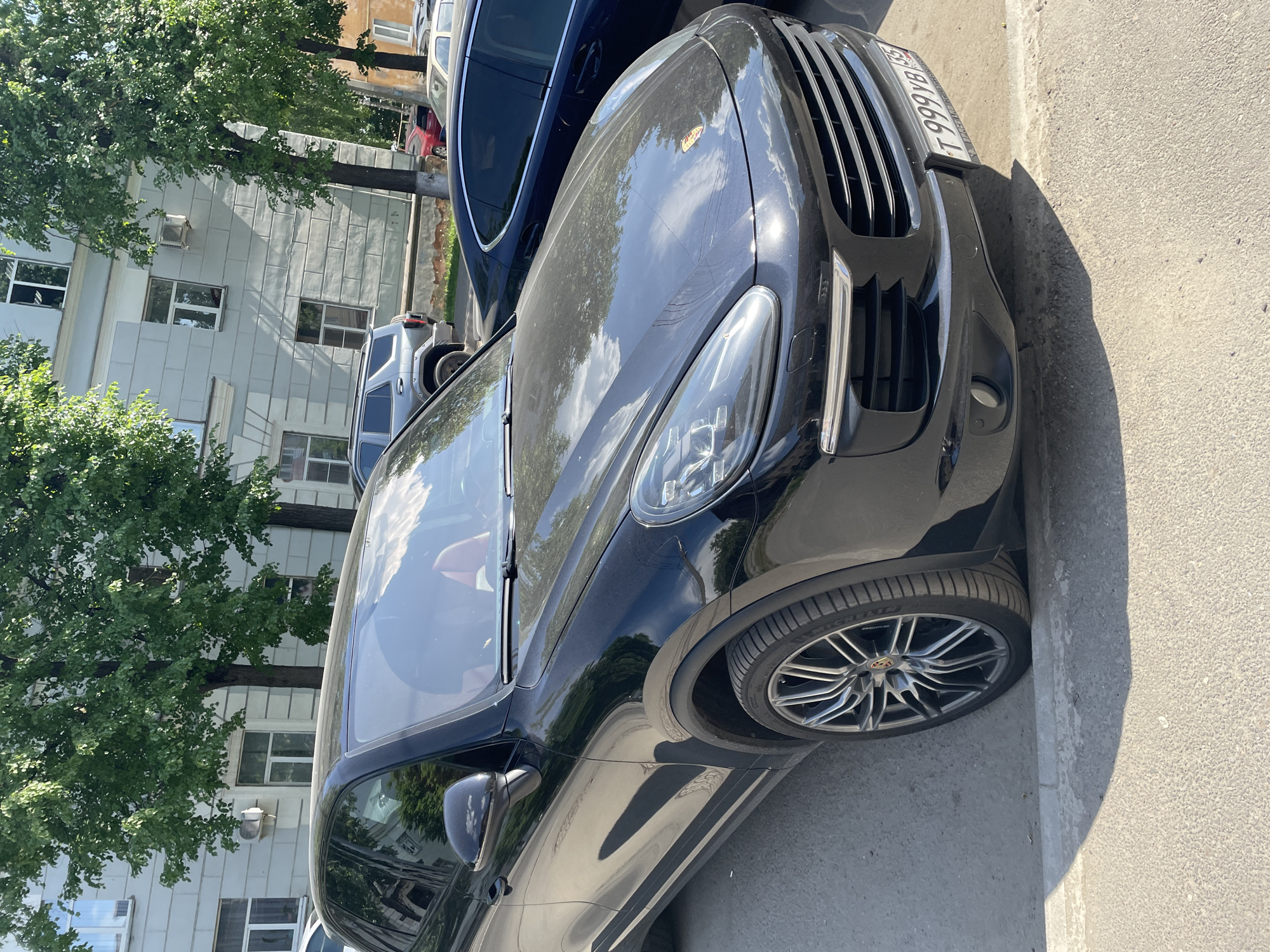 The image shows a black Porsche Cayenne parked on a street in Vologda, Russia. The car is positioned in the foreground, angled towards the viewer. The building behind the car appears to be a light-colored, multi-story structure with several windows. Trees with green foliage are visible, casting shadows on the car and the pavement. The license plate of the Porsche is partially visible and reads "T 999 YB 35". The scene is illuminated by sunlight, suggesting it is daytime.