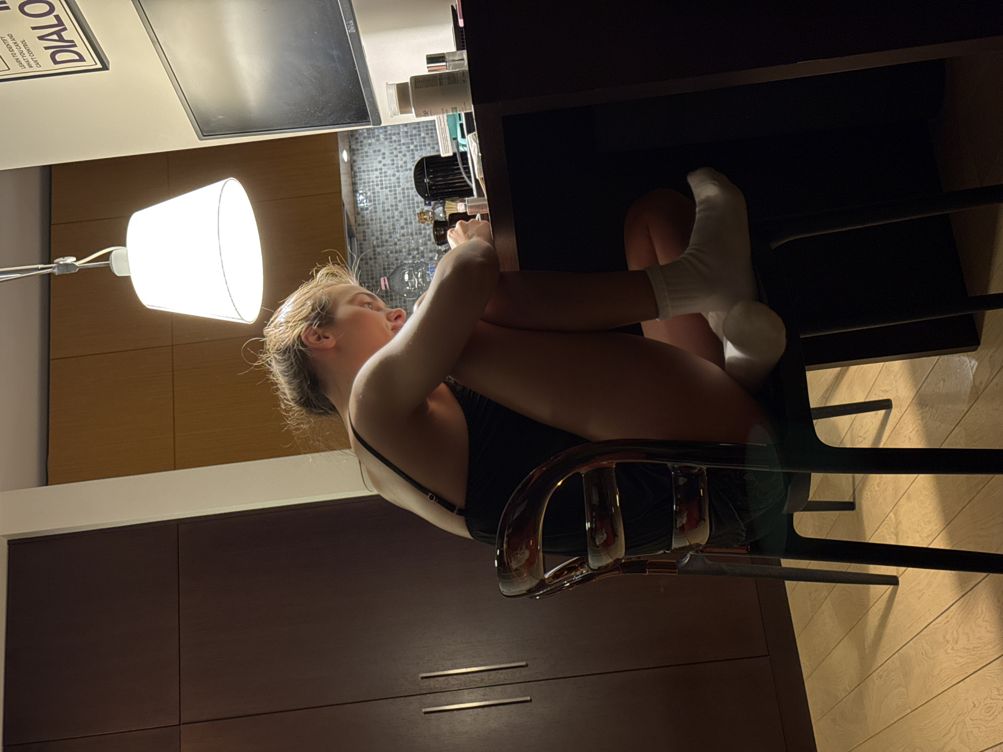 A young woman is seated on a chrome-backed chair, drawn up to a dark-colored desk. She is wearing white socks and a dark-colored top. Her knees are pulled up to her chest, with her arms wrapped around them. The desk has a small selection of toiletries and a beverage on it. A bright lamp is overhead. The location appears to be an apartment or hotel room in Warsaw, Poland. This is suggested by a sign on the wall. Dark wood cabinetry is visible. The floor is wood.