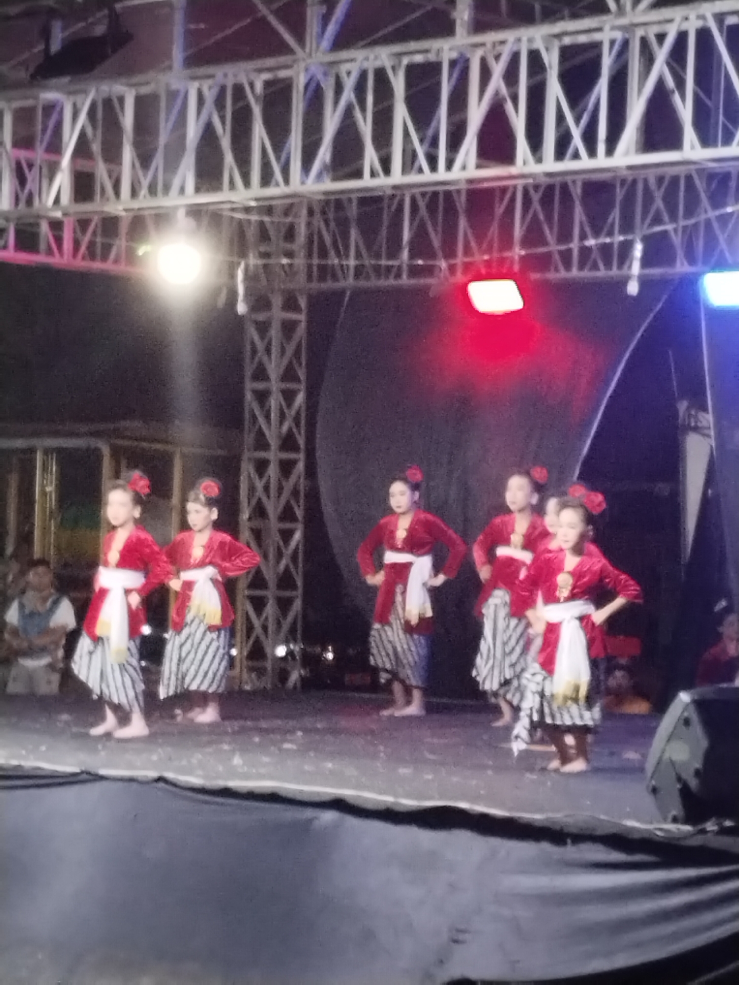 The image depicts a group of young girls performing a traditional dance on an outdoor stage in Kecamatan Pati, Indonesia. The girls are dressed in elaborate red and white traditional costumes, with their hair adorned with red flowers. They are all barefoot and appear to be moving in unison. The stage is lit by a series of lights, with a red light prominently visible at the top center of the frame. In the background, there are spectators, some of whom are visible in the cropped images. The overall atmosphere suggests a cultural event or performance.
