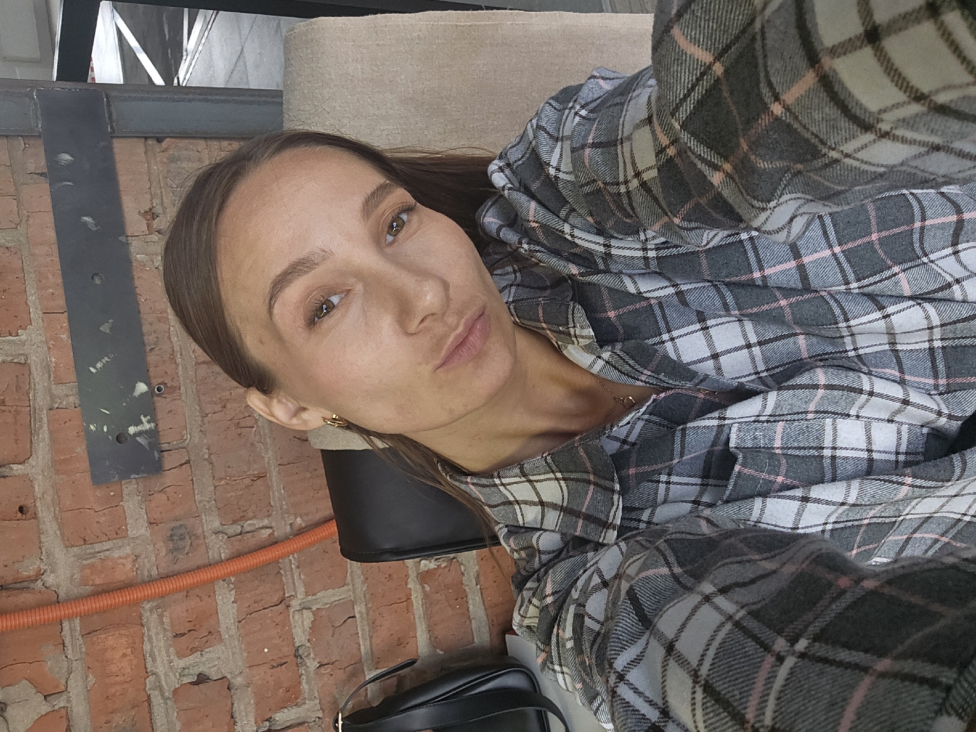 A woman is lying down, facing the camera. She has long brown hair and is wearing a gray, white, and pink plaid shirt. A black leather bag is visible below her. Behind her is a brick wall with an orange pipe running horizontally along it. A black metal structure is also visible against the wall. The location is Chaikovsky, Russia.