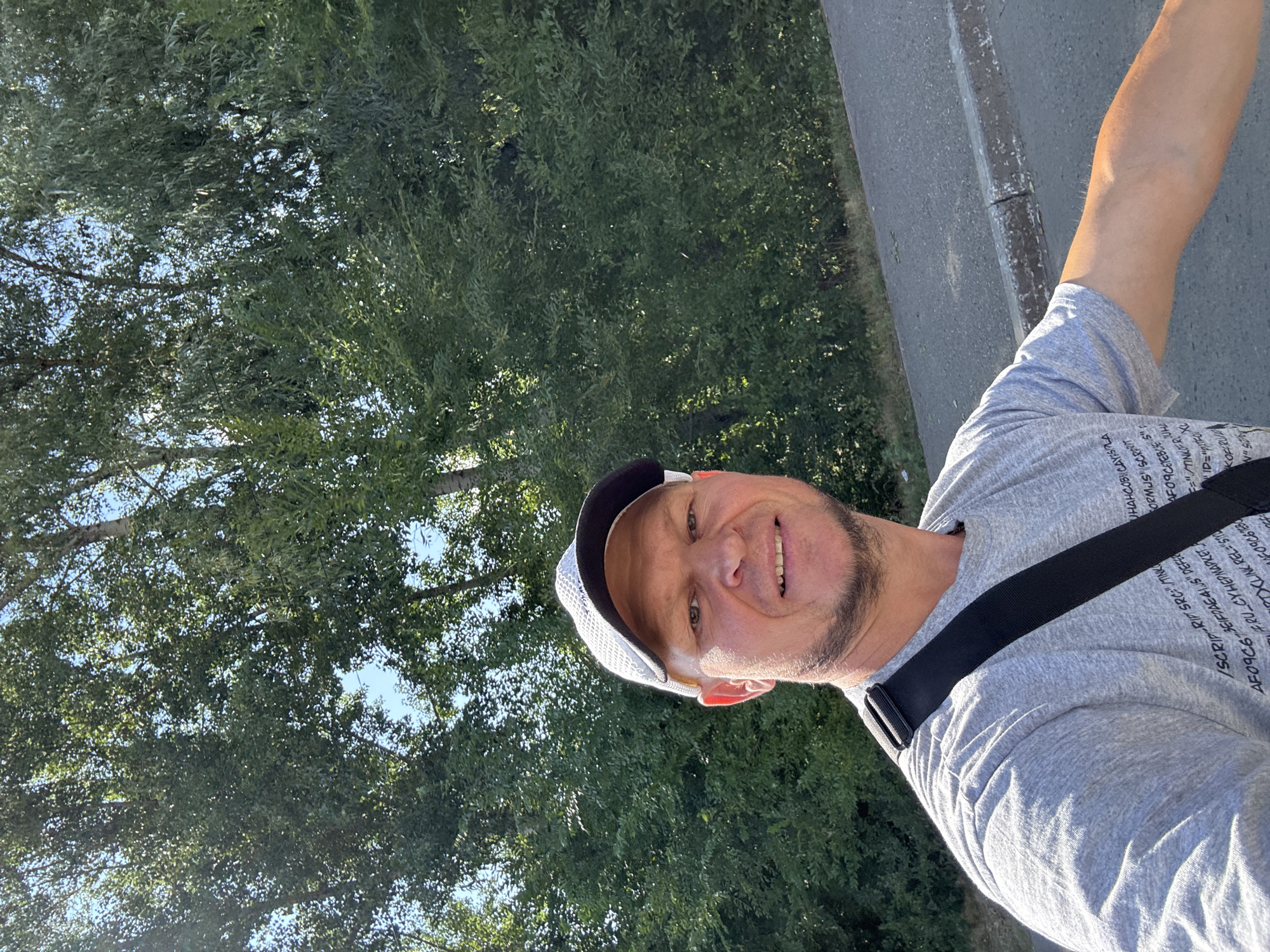 A man is taking a selfie outdoors on a sunny day. He is wearing a grey t-shirt, a white and black baseball cap, and a black crossbody bag. He has short hair and a beard, and he is smiling. The background is filled with green trees and a blue sky. A paved road and a curb are visible in the lower right corner of the image. There are no specific landmarks visible to identify the city of Oskemen, Kazakhstan.