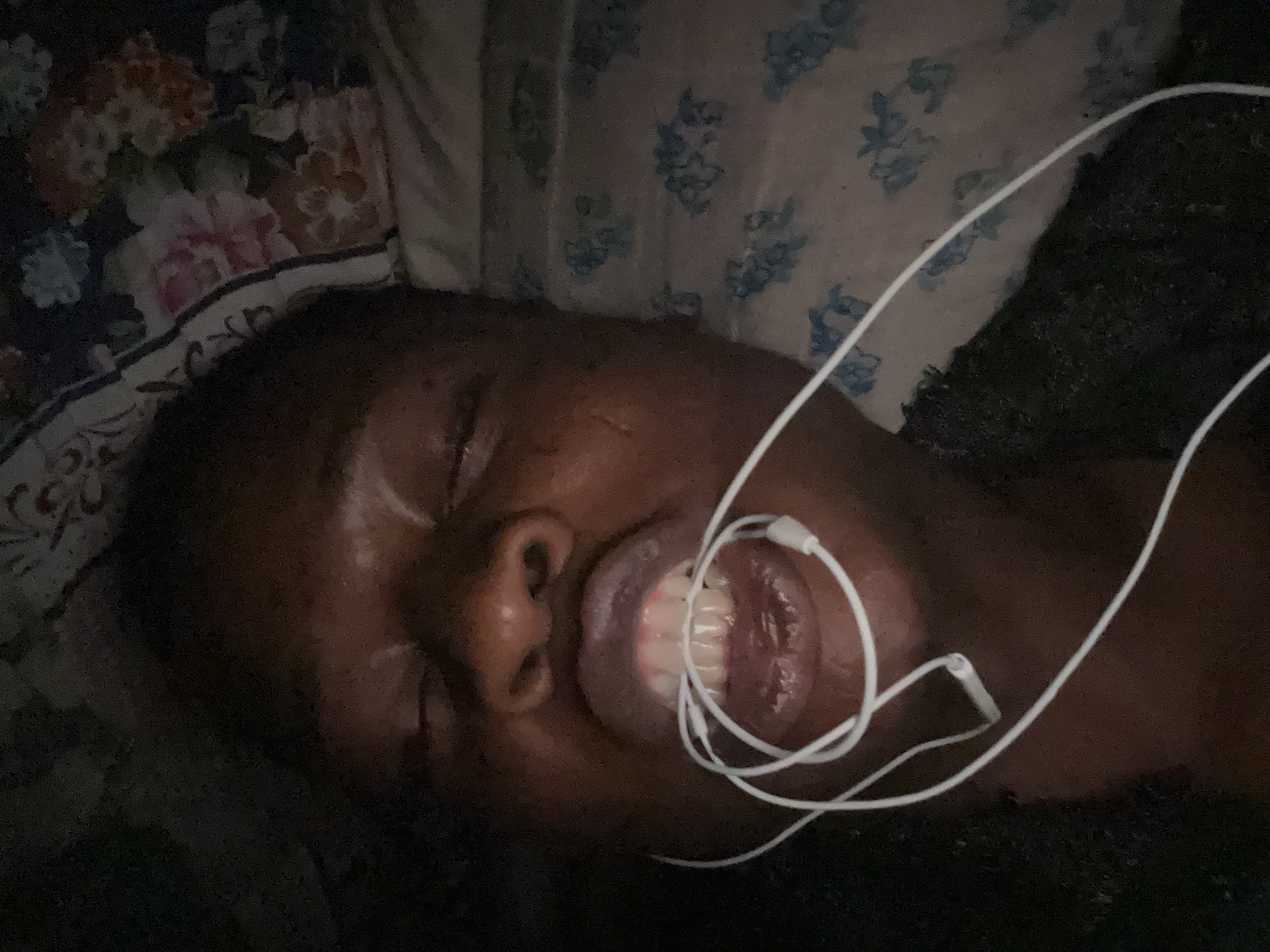 A person with dark skin lies on a pillow with a floral print, eyes closed and mouth slightly open. White earbud headphones are draped across their face and inside their mouth. The background is dark.