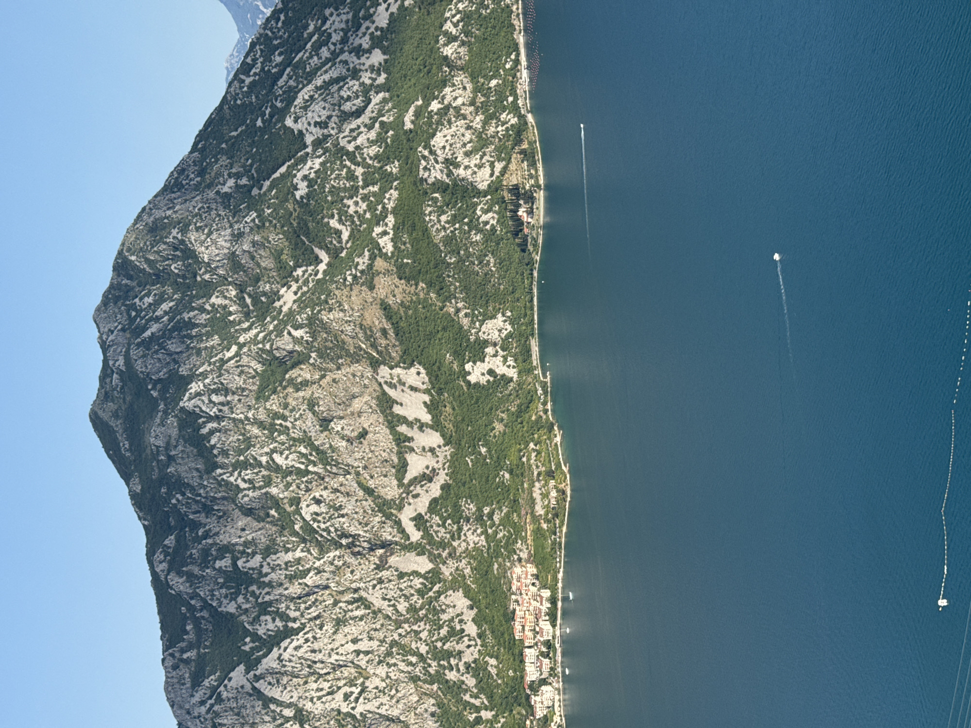 This is a vertical, eye-level, medium shot of a coastal town nestled at the base of a steep, rocky mountain range overlooking a wide expanse of deep blue water. The mountain dominates the background, its rugged slopes dotted with green vegetation and crisscrossed by light-colored rock formations. In the middle ground, a cluster of buildings with red-tiled roofs forms a small town situated along the shoreline. Several sailboats are visible near the shore. The water in the foreground is calm, with a single white boat leaving a faint wake. The sky is clear and blue. The scene appears to be the town of Štrp, Montenegro, as indicated by the distinctive mountain and coastal landscape.