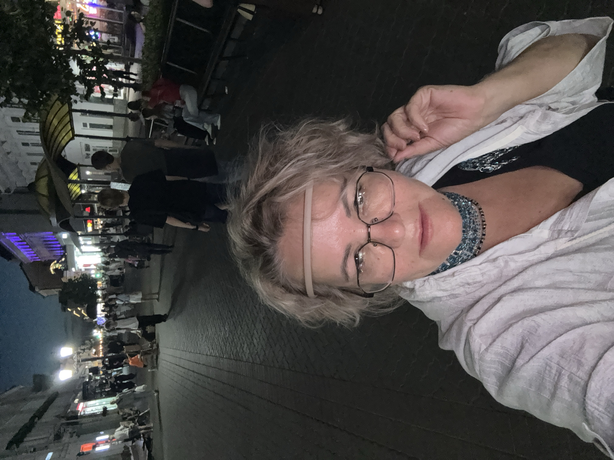 A full shot taken at night, at eye level, shows a woman with blonde hair wearing glasses and a light-colored jacket. The woman is holding her hair with her right hand. She is wearing a necklace that appears to be made of blue and black beads. She is standing in a paved walkway, with shops and people in the background. The scene suggests a street in Kazan, Russia, possibly during a festival or event, with lights from shops and street lamps illuminating the area. The background shows a crowd of people walking and standing, some sitting on benches. The overall atmosphere is lively and urban.