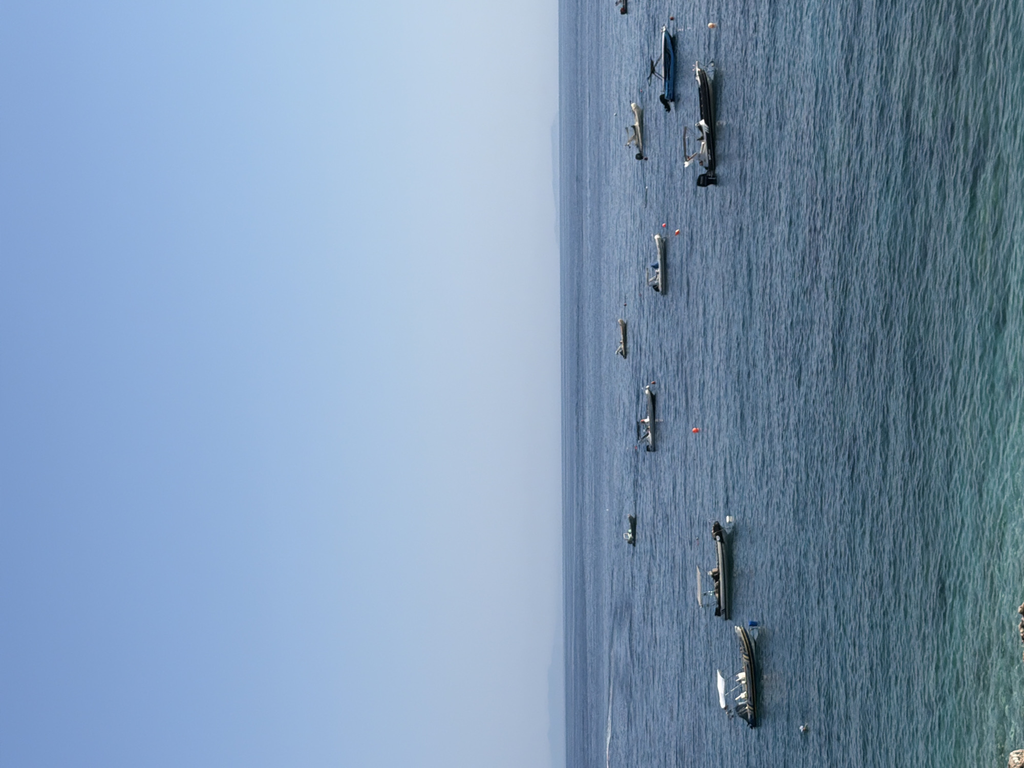 The image depicts a calm sea scene with multiple small boats anchored in the water. The sky is a clear, pale blue, and the water is a deep blue, with some lighter turquoise hues visible in the foreground, suggesting a shallow area or a rocky seabed beneath the surface. Several white and grey motorboats are scattered across the frame, some with canopies. In the distance, a hazy line of land is visible on the horizon. The overall impression is one of a serene coastal setting. This scene is likely from the city of Himarë, Albania, a coastal town known for its beautiful beaches and clear waters.