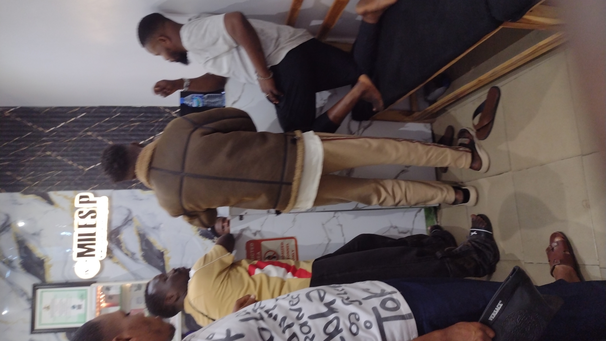 The image shows a group of men in an interior space. The location is in the city of Unknown, Unknown. The setting features a decorative wall with gold marble patterns and a lighted sign that reads "MILESP."
There are four men visible in the image. One man wears a white T-shirt and black pants and stands near the wall. Another man is seen in a brown jacket with light-colored pants. Further down, a man wears a yellow shirt with a patterned white shirt on top. Another man near the bottom of the image is dressed in a dark blue shirt with a white patterned T-shirt.
Several pairs of shoes are visible on the tiled floor. There is a "No Smoking" sign on the wall. A water bottle is also present.
The men seem to be in a waiting area or lounge. One man is seated on a wooden bench. Some interactions seem to be occurring, but the exact nature is unclear.