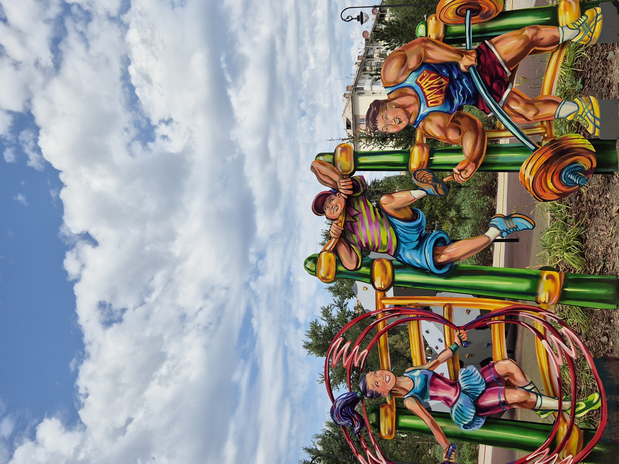 This is a vertical, medium shot of a colorful outdoor art installation on a playground, likely in Kokshetau, Kazakhstan, under a partly cloudy blue sky. The installation features three cartoonish, larger-than-life figures engaged in athletic activities. On the left, a young girl with dark purple hair in a ponytail is depicted jumping rope. She wears a turquoise tank top, a pink and blue skirt, and green sneakers. Her right arm is extended, holding a jump rope that forms a red circular motion around her. In the center, a boy wearing a striped shirt with pink, yellow, green, and purple stripes, and blue shorts, is shown on a climbing structure, with his right arm gripping a green bar and his left hand resting on his head. To the right, a muscular man in a blue tank top with a red "QAMAR" logo and red shorts is lifting a barbell with large, colorful weights. He wears yellow sneakers and his left arm is raised with his index finger pointing upwards. The art is mounted on green and yellow supports, with some areas of greenery and wood chips visible at the base. In the background, trees and residential buildings are visible, along with a street lamp.