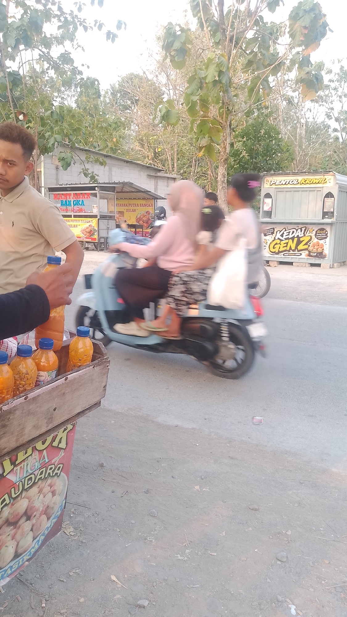 In Karanglangu, Indonesia, an outdoor scene unfolds with a focus on a roadside vendor and passing traffic. A man in a light-colored polo shirt stands on the left, with his right arm extended towards a wooden crate filled with orange bottles, some of which have blue caps. The crate also features a sign with red and yellow lettering, with the words "TIGA" and "AUDARA" partially visible, along with an image of round food items.
In the mid-ground, a light blue scooter carries two individuals. The person in front wears a pink headscarf and a light-colored top, while the person behind them wears a dark shirt. Both are blurred due to motion. A white bag is visible on the back of the scooter.
In the background, several structures are visible. To the left, a building with a gray roof and white walls displays a sign that reads "MARTABAK BANGKA" and other text. To the right, a white food truck or stall has "PENTOL KRIWIL" written on it, and another sign reads "KEDAI GEN Z" with an image of fried food. Trees with green foliage line the background, suggesting a semi-rural or suburban setting. The ground is paved with a rough, grey surface, and scattered debris is present. The overall lighting suggests it is daytime, possibly late afternoon, given the warm tones.