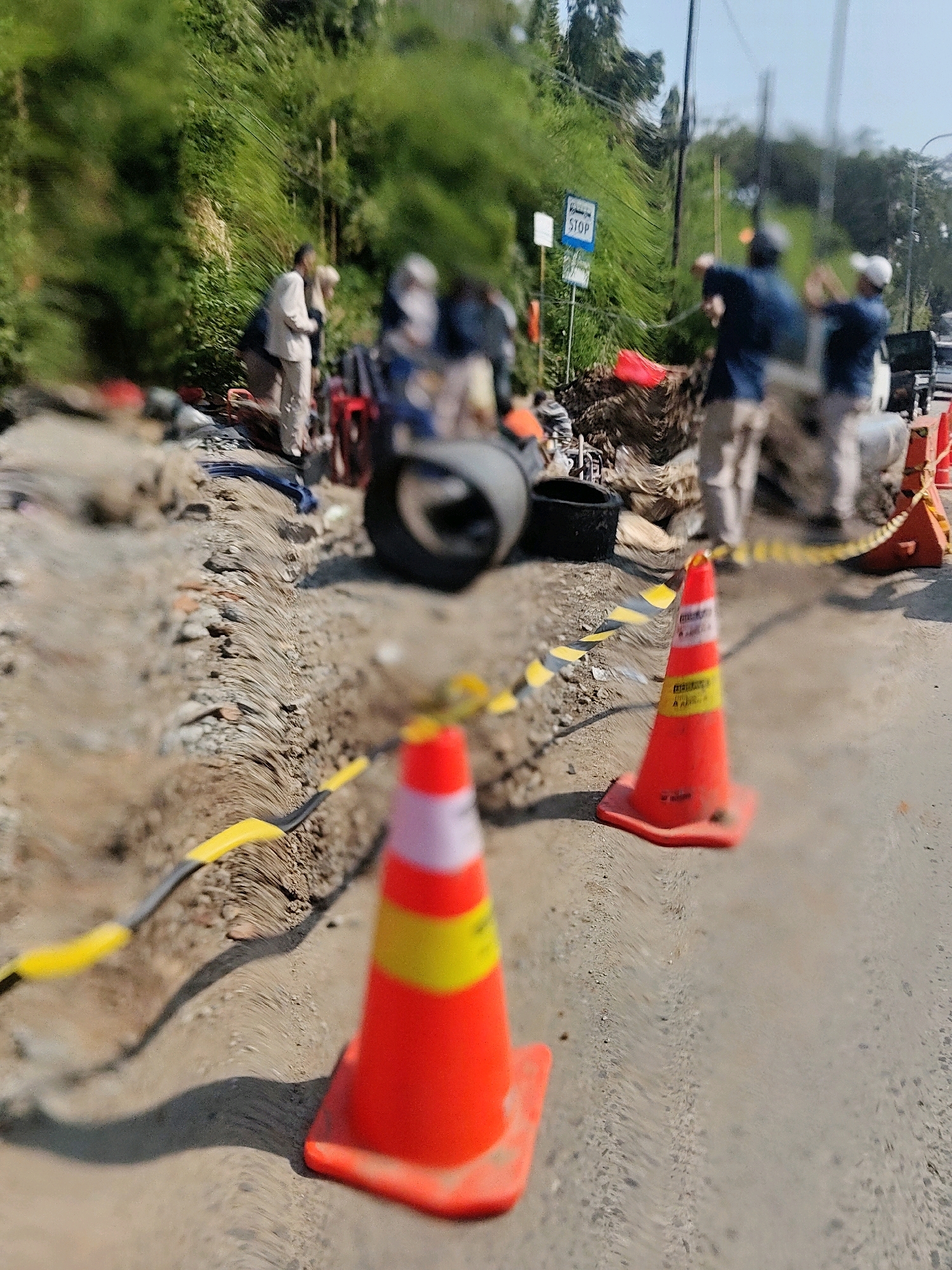 The image depicts a street scene in Pasirsari, Indonesia, with a visible road under construction or repair. Several orange traffic cones, some with reflective yellow and white bands, are strategically placed to direct traffic or mark a hazard. A black and yellow striped caution tape is also visible, reinforcing the demarcation of the construction area. In the background, several people appear to be involved in the work or observing the situation. One person is wearing a light-colored jacket and pants, while others are in darker clothing. A blue sign with the word "STOP" is present on a pole, indicating a traffic control measure. Debris, including dirt, rocks, and what appears to be a tire, are scattered around the construction site. The overall environment suggests an active roadwork operation in a semi-urban or rural setting, characterized by lush green vegetation along the roadside.