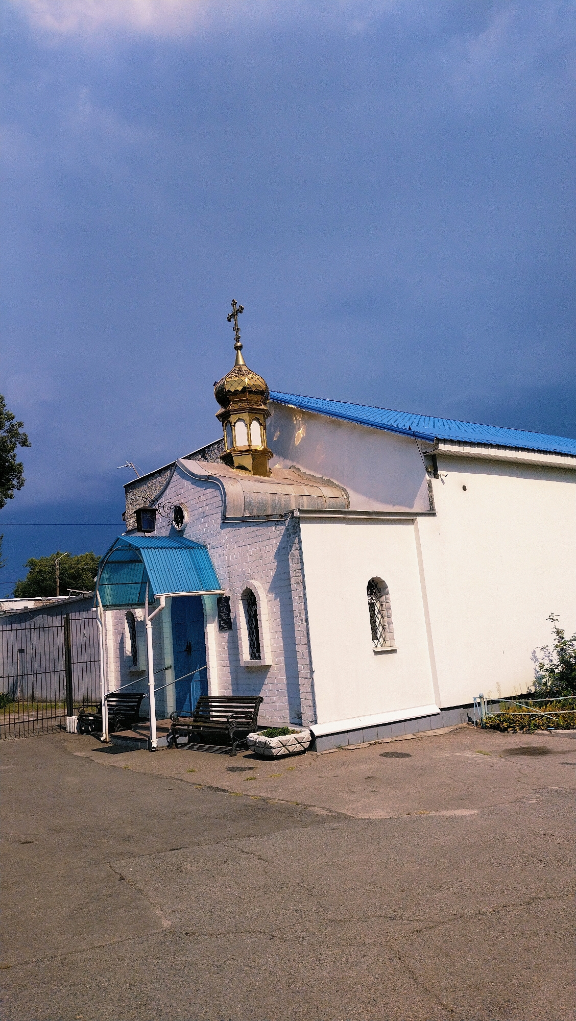 The image shows a small, white-brick church with a blue metal roof in Horishni Plavni, Ukraine. The church features a golden dome topped with a cross, and a blue awning over the entrance. Two dark-colored benches are placed in front of the church, with a planter box to the right. A metal fence and gate are visible to the left of the church. The sky is a deep blue with dramatic, dark clouds. The ground in front of the church is paved with asphalt. There are no people visible in the image.