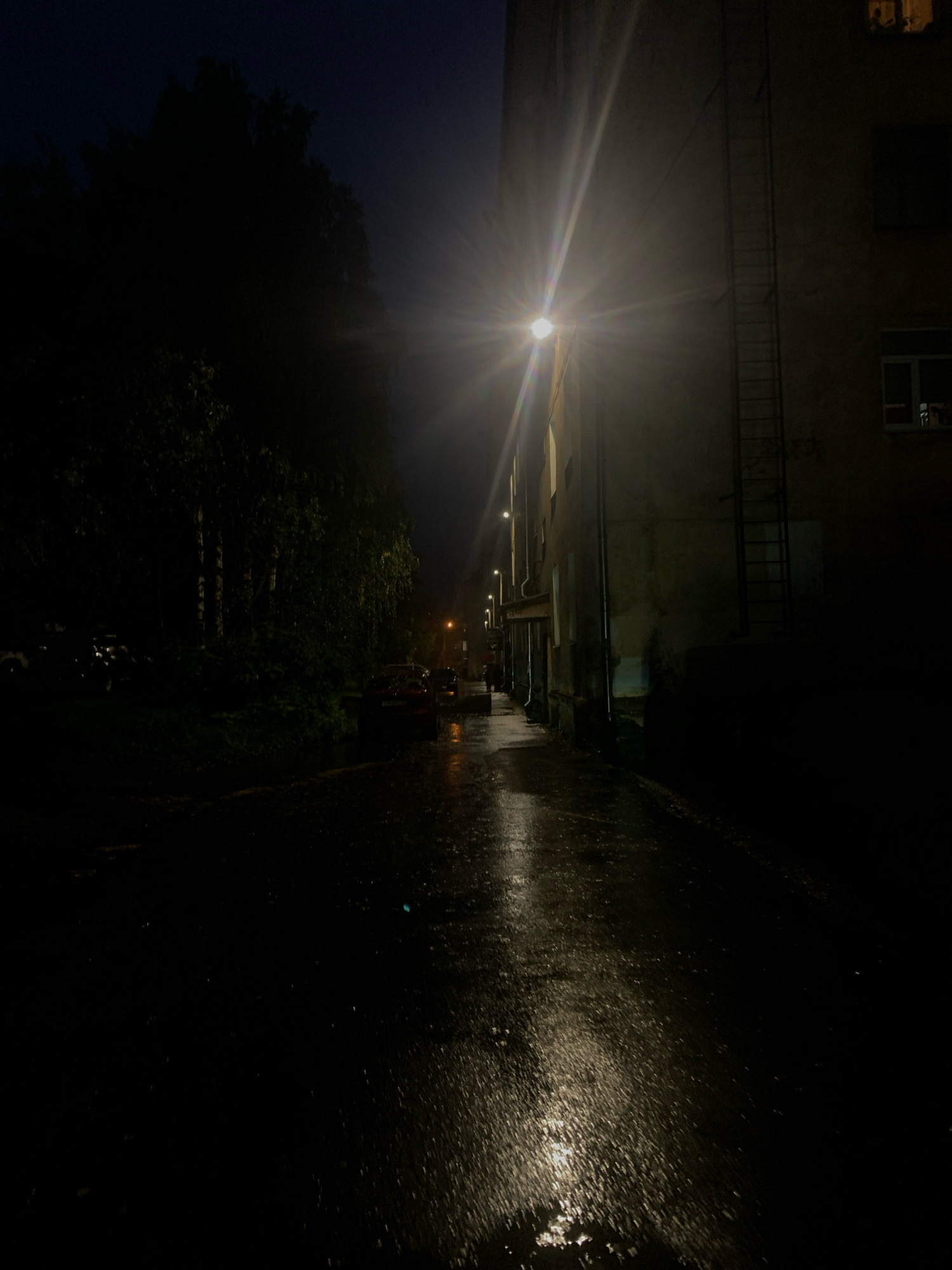 The image depicts a dimly lit street scene at night in Arkhangelsk, Russia, likely a residential area given the presence of a multi-story building and parked cars. A bright street lamp illuminates the wet asphalt, creating a reflection that stretches down the street. Trees line the left side of the street, their dark foliage contrasting with the illuminated building. The building itself appears somewhat aged, with a fire escape visible on its side and windows that show faint interior light. Several cars are parked along the street, some of which are partially obscured by the darkness and the foliage. The overall atmosphere is one of quiet, nighttime stillness, with the wet pavement suggesting recent rain.
