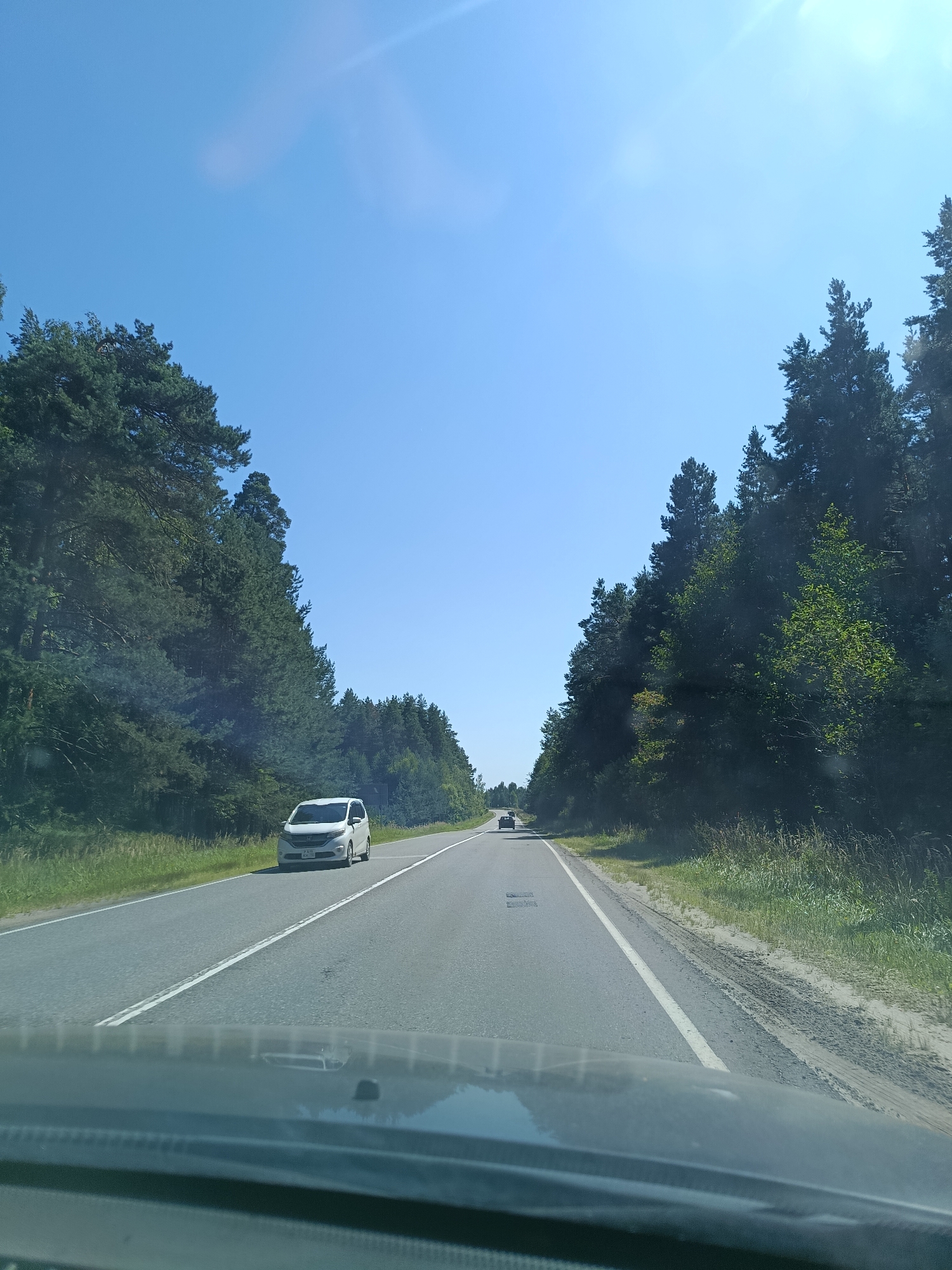 The image depicts a view from inside a vehicle driving on a two-lane asphalt road lined with dense coniferous forest on both sides. The sky is clear and blue with a hint of sun glare. A white minivan is visible on the right lane of the road, moving away from the camera, and another darker vehicle is further ahead on the left lane. The road has white lane markings. The foreground shows the dashboard and windshield of the vehicle from which the photo is taken. The environment suggests a rural or suburban setting in a wooded area, possibly along a highway or main road. The location is identified as being in the city of Savino, Russia.