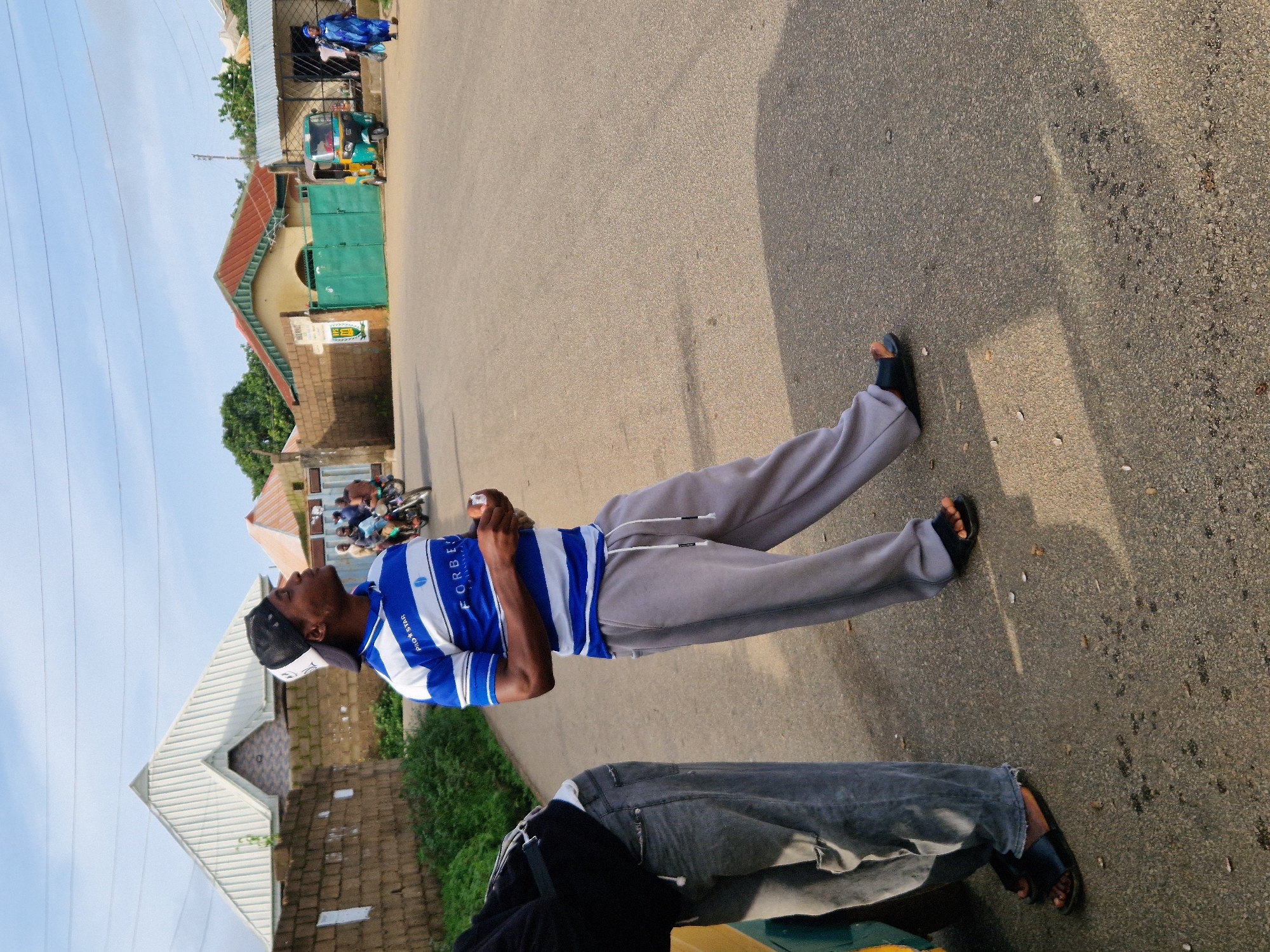 The image depicts a street scene in Kabong, Nigeria, under a partly cloudy sky. In the foreground, a Black man wearing a blue and white striped polo shirt with "FORBE" visible on it, grey sweatpants, a black baseball cap, and black slides, stands looking upwards and to his right. He is holding a small white object in his hands. To his left, another person is partially visible, wearing a black jacket and grey jeans, leaning against a yellow object, likely a vehicle.
The background shows a street with a paved surface. On the right side of the street, a green gate is prominent, with a yellow and green auto-rickshaw parked in front of it. A woman in a blue dress is walking away from the auto-rickshaw towards the right edge of the frame. Behind the gate, a building with a red roof and light-colored walls is visible, along with trees and other structures. On the left side of the street, a brick wall runs along the roadside, with a large, modern building with a white, pyramidal roof behind it. The overall impression is of a bright, sunny day in a residential or commercial area.