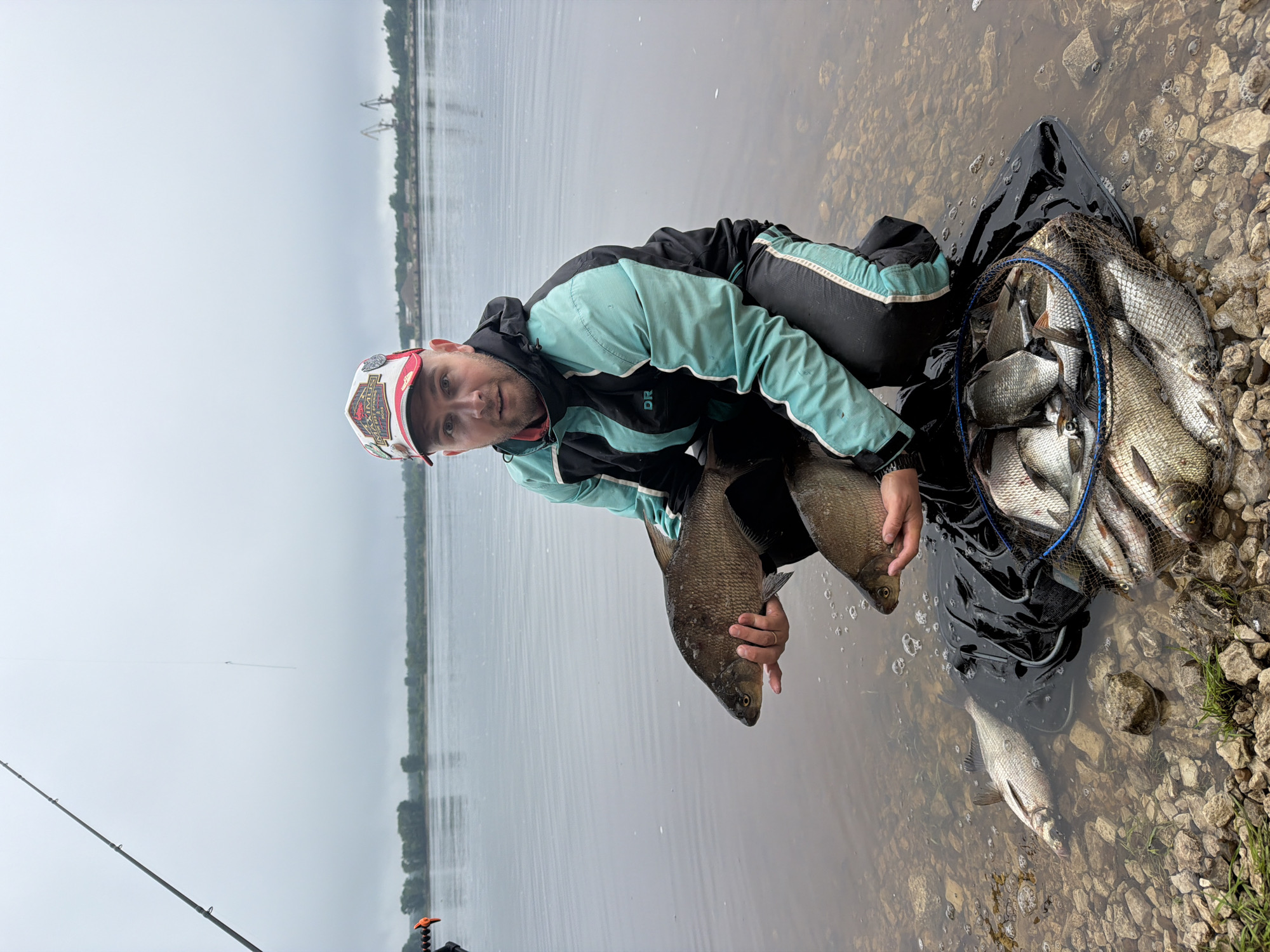 The image captures a fisherman showcasing his catch on an overcast day by a large body of water in Chaikovsky, Russia. The central figure is a man, seen crouching in the shallow water near a rocky bank. He is wearing a black and teal jacket with "DR" visible on the sleeve, black pants with white stripes, and a white and red baseball cap emblazoned with "MAXIMUS" and "WORLD TOUR." He holds two substantial, silvery-brown fish (likely bream or similar freshwater species) in his hands, looking towards the viewer with a neutral, possibly contented expression.
Adjacent to the man, a black keepnet with a blue mesh rim is partially submerged in the water, brimming with numerous other fish, indicating a significant catch. One fish is also visible lying in the clear, shallow water among the rocks and pebbles. A fishing rod, set up with a stand, extends into the lower left corner of the frame, further emphasizing the activity.
The setting features a wide expanse of calm, grey water under a cloudy sky. The far bank is dimly visible in the distance, lined with trees and what appear to be a few industrial structures or cranes, adding a hint of urban landscape to the natural scene. The immediate foreground consists of a rocky and pebbly shoreline where the man is situated. The overall impression is one of a successful fishing excursion on a quiet, possibly cool, day.