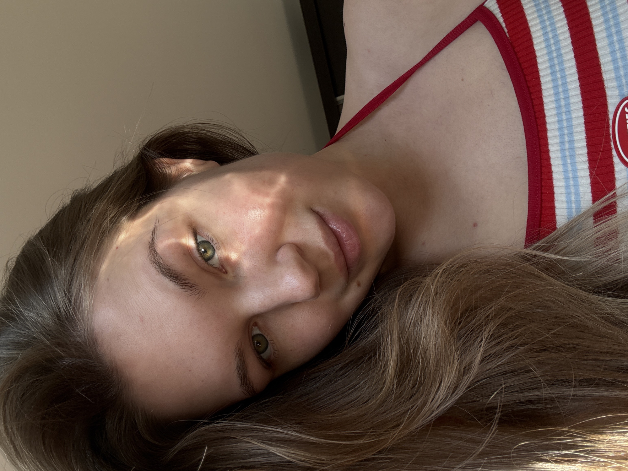 A high-angle, close-up portrait captures a fair-skinned individual, likely a young woman, lying down and looking directly at the viewer. The image is oriented 90 degrees clockwise, presenting the person's head tilted to the right. She has striking green eyes, dark eyebrows, and long, wavy hair that blends brown and blonde tones, spread around her head. Her lips are naturally parted. Sunlight streams across her face, creating distinct patterns of light and shadow, suggesting a specific time of day, possibly morning or late afternoon.
She is wearing a ribbed top featuring bold red and white stripes, interspersed with thinner light blue stripes. A red strap of the top is visible over her shoulder. On the upper right side of her chest, a circular patch on the garment is partially visible, showing the letters "RIES". The background is a plain, uniformly lit light beige/tan color, indicative of an indoor setting. Her expression is calm and neutral. No specific location cues related to Vozdovac Urban Municipality, Serbia, are visible within the image.