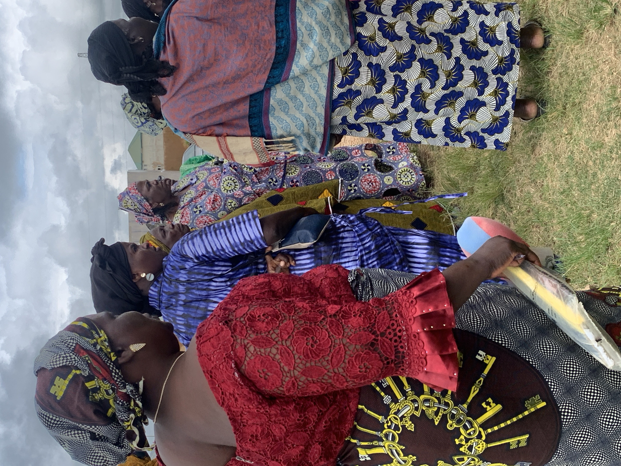 The image shows a group of women standing outdoors, possibly at an event or gathering, with a cloudy sky overhead. The women are dressed in brightly colored, patterned African attire. The woman in the foreground on the left wears a red lace top with ruffled sleeves and a patterned skirt featuring a large key design. Next to her, another woman wears a blue and white tie-dye-style outfit. Further back, other women are visible in various patterned dresses and headscarves. In the background, there is a glimpse of buildings and what appears to be some industrial infrastructure, possibly hinting at a semi-urban or peri-urban setting within the city of Jos, Nigeria. The ground is covered in grass. One woman is holding a rolled-up object in plastic packaging.