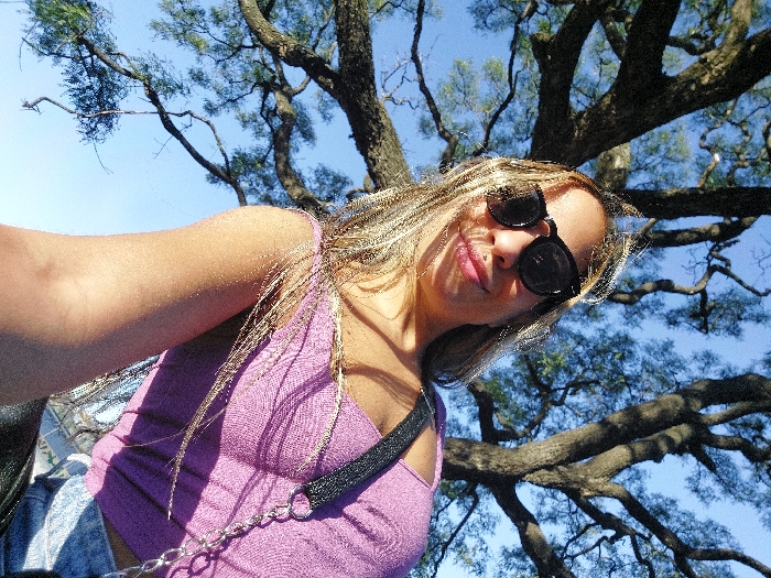 A female individual is depicted in a low-angle outdoor selfie against a clear blue sky and the canopy of a large, mature tree featuring thick, gnarled branches and sparse green leaves. The subject wears dark sunglasses, obscuring her eyes, and has a small stud piercing in her left nostril. Her lips are visible, colored light pink and slightly parted. She possesses long, light-colored hair, appearing somewhat windswept, falling around her face. She is dressed in a light purple, possibly ribbed, crop top, with a black strap visible over her left shoulder and a silver chain accessory near her upper torso. A fragment of a denim garment is visible at the bottom right of the frame. The scene is illuminated by direct sunlight, casting subtle shadows on the subject. Based on the characteristics of the large tree and outdoor park-like setting, the location is consistent with a public green space within the city of Buenos Aires, such as the Palermo Parks.