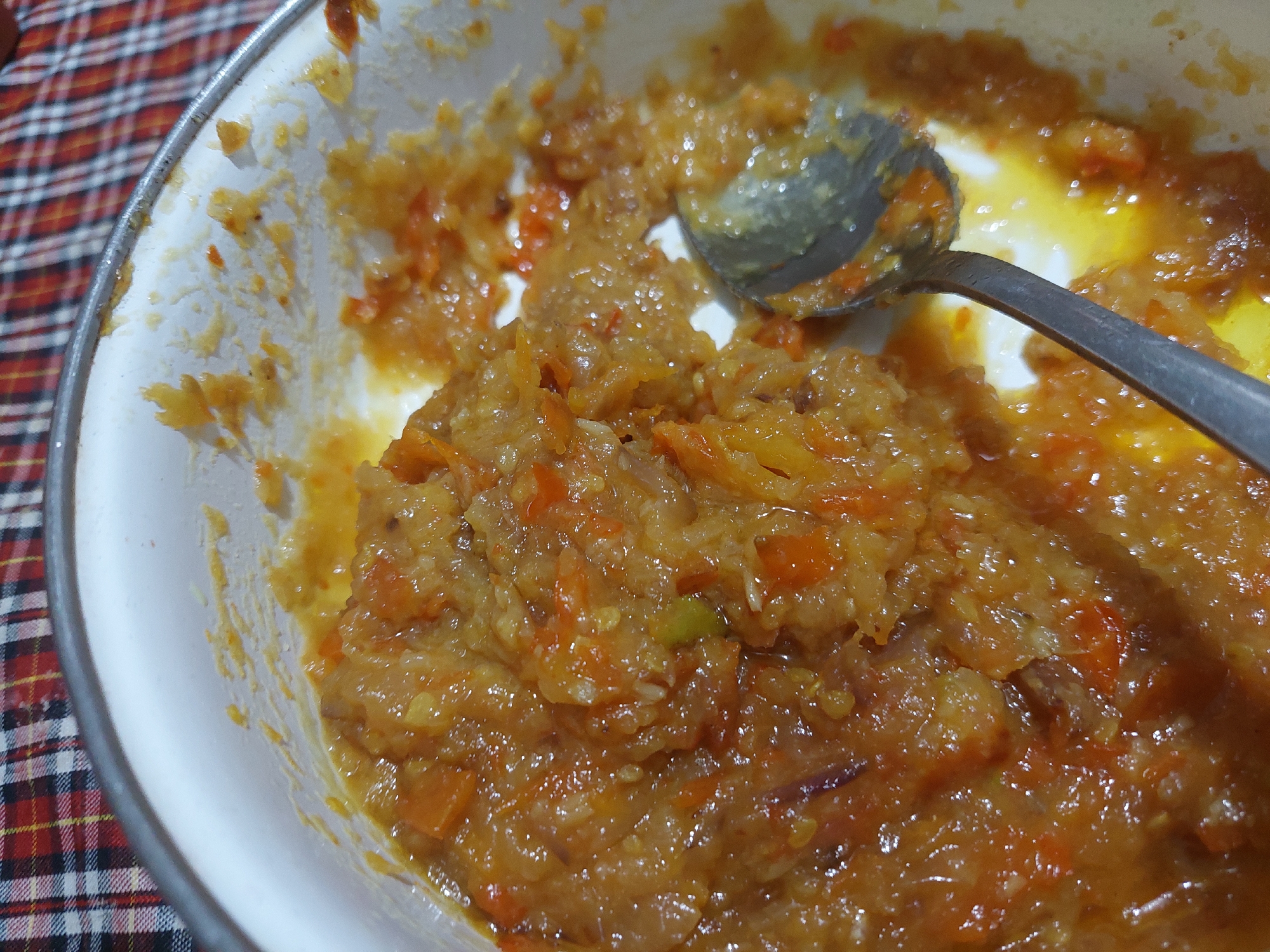 A close-up, overhead shot shows a serving of cooked shredded vegetables, likely carrots and possibly other root vegetables, in a white bowl. The vegetables are finely shredded and appear to be stewed, with a glistening, moist texture and an orange-brown hue. A metal spoon rests in the bowl, scooping a portion of the mixture. The bowl is placed on a red and black checkered tablecloth. The scene is set indoors, potentially in a kitchen or dining area in Jelutung, Indonesia, suggested by the checkered pattern often used in Indonesian households.