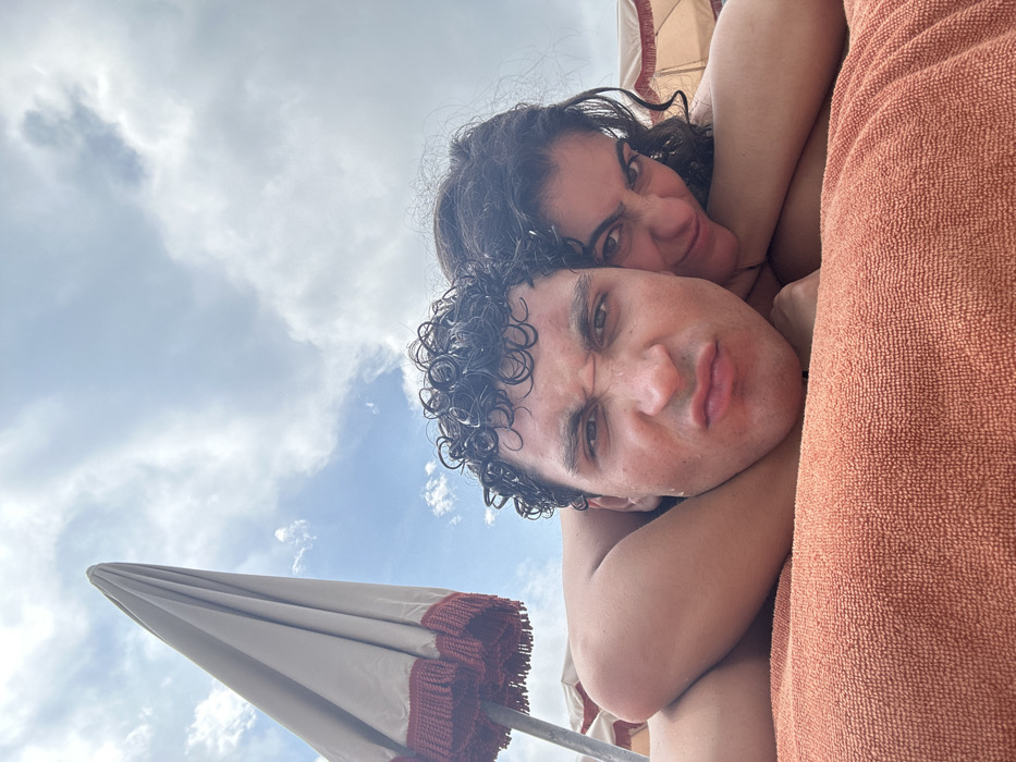 A low-angle shot captures a couple against a backdrop of clouds and blue sky. The man in the foreground, with wet curly hair and a slightly scowling expression, is partially obscured by a textured orange towel. A woman behind him, also with curly hair, grins into the camera. An umbrella's pointed top, trimmed with dark red fringe, is visible in the lower left. The scene is brightly lit, suggesting an outdoor, possibly beachside, location.