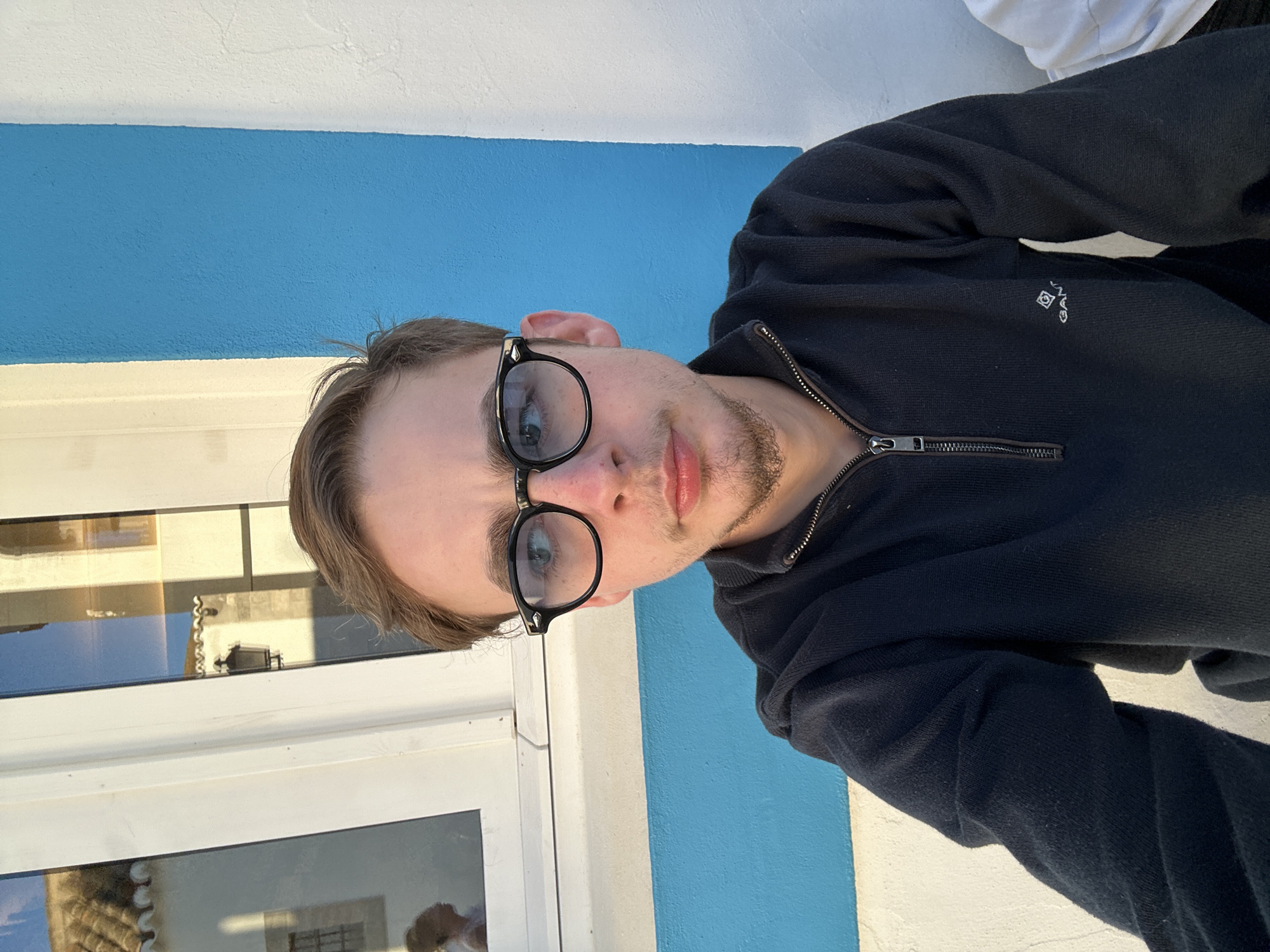 The image is a selfie of a young man wearing black-rimmed glasses and a dark blue zippered sweater. The sweater has a small white logo on the left side of the chest. He appears to be sitting outdoors near a window with white frames and blue walls, possibly in Vila Real de Santo António, Portugal. The window reflects an image of what seems to be buildings or a townscape.