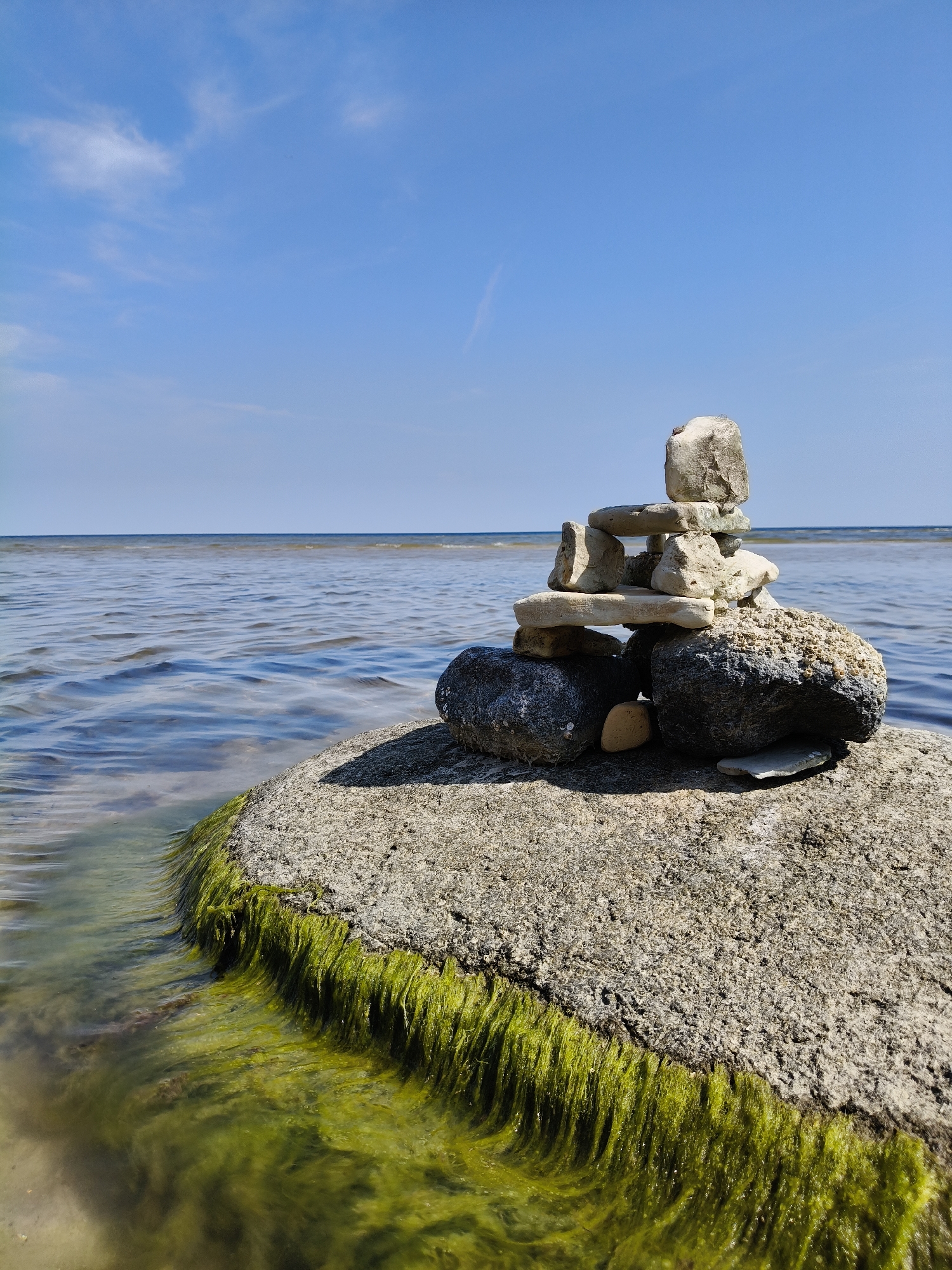 A balanced rock formation, resembling an inukshuk, is centrally positioned on a large, rough-textured boulder. The boulder is partially submerged in clear, calm water, with vibrant green algae clinging to its submerged surface. Behind the boulder and the rock formation, the horizon stretches across a vast expanse of water, meeting a clear blue sky dotted with wispy white clouds. The overall scene evokes a sense of tranquility and natural artistry, situated on the shores of Bigauņciems, Latvia.