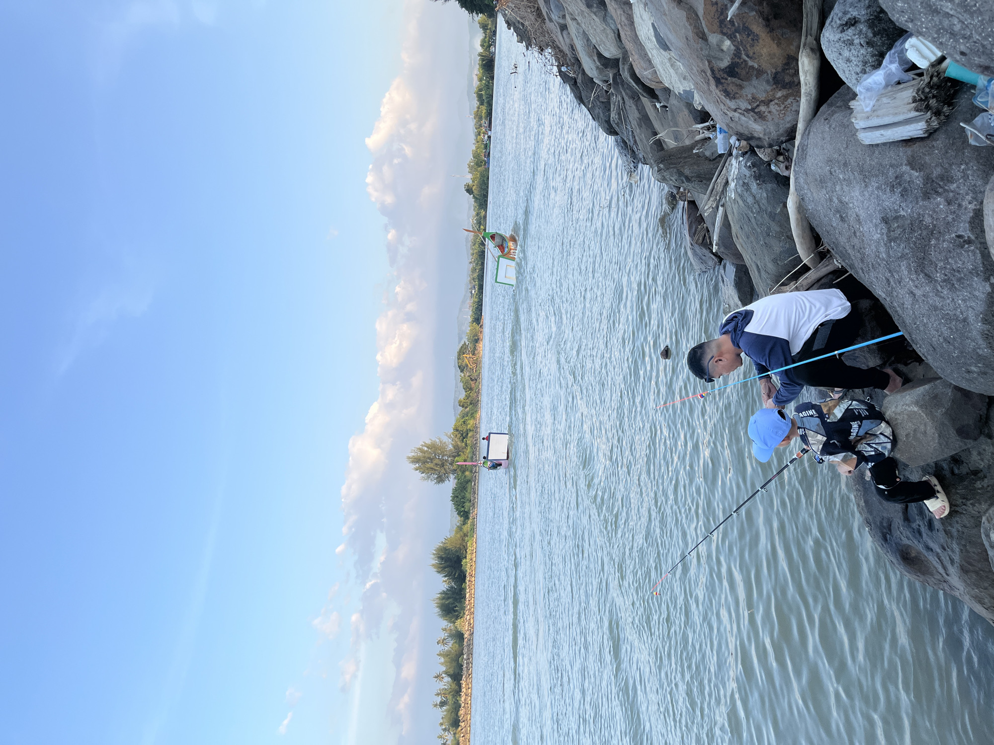 In the city of Reudeuep, Indonesia, two individuals, an adult male and a young boy, are fishing from a rocky shore. The boy, wearing a blue cap and a camouflage-patterned shirt, sits on a large rock, holding a fishing rod. Beside him, the man, dressed in a white and blue hooded jacket, also holds a fishing rod, appearing to assist or teach the boy. The water is calm and reflects the blue sky dotted with white clouds. In the distance, two small boats are visible on the water, and a treeline borders the opposite shore. The foreground is dominated by large, grey rocks, some with driftwood and plastic debris scattered among them.